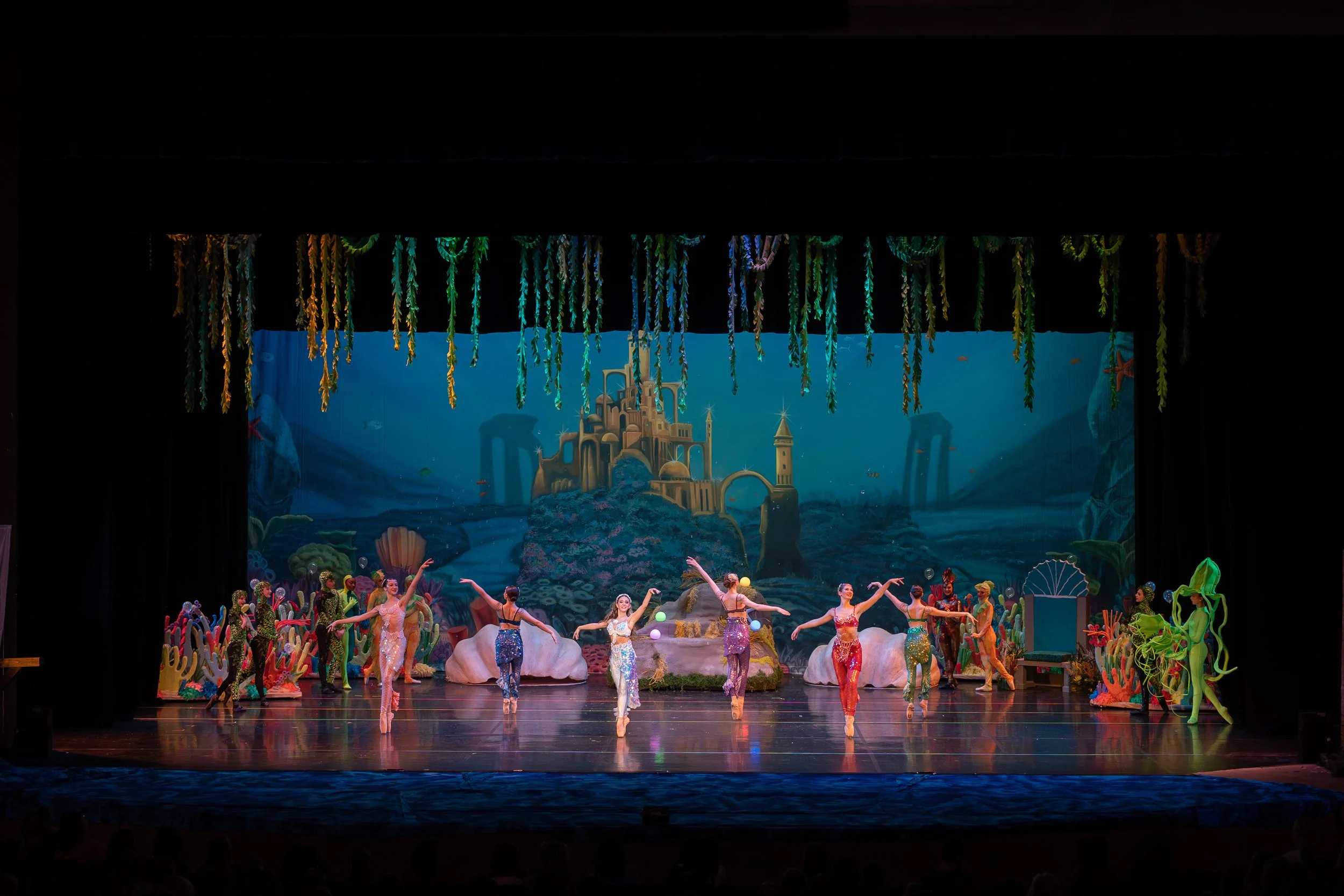 Colorful stage performance of a ballet or dance show inspired by Under the Sea theme, with dancers in vibrant costumes and a scenic backdrop of an underwater castle and ocean scene.