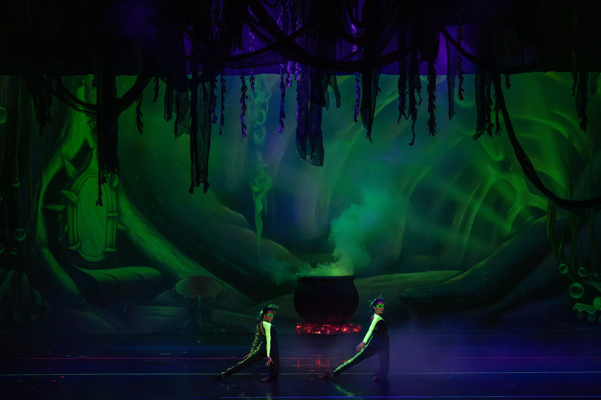 Two performers in black and green costumes with visors perform a dance on stage in a dark, theatrical setting with a mystical forest backdrop. A large cauldron or pot with green smoke rises above it in the background, creating a fantasy or magical at