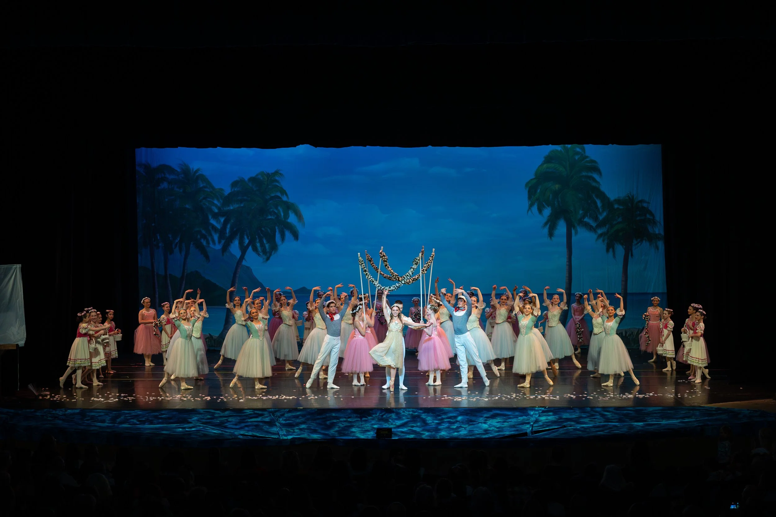 Ballet dancers on stage performing under a tropical backdrop with palm trees and blue sky, with some dancers in pink tutus and others in pastel dresses, during a scenic production.