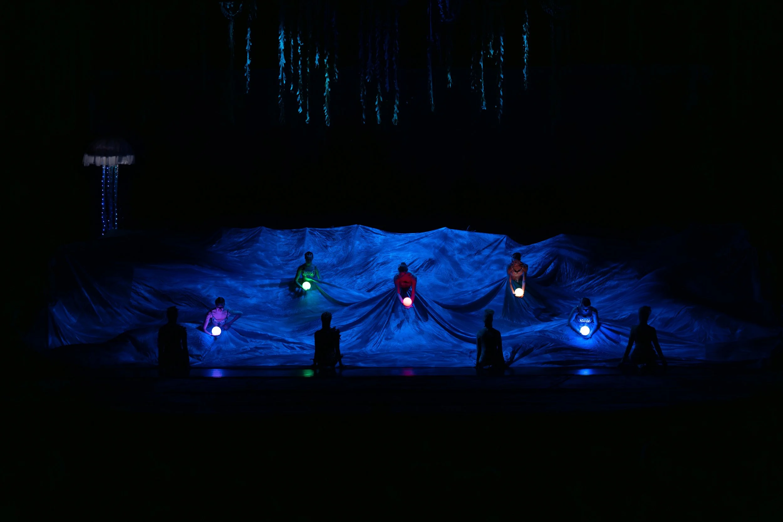 A stage performance with six performers holding glowing orbs, beneath hanging decorations resembling jellyfish, with a water-like textured backdrop illuminated in blue light.
