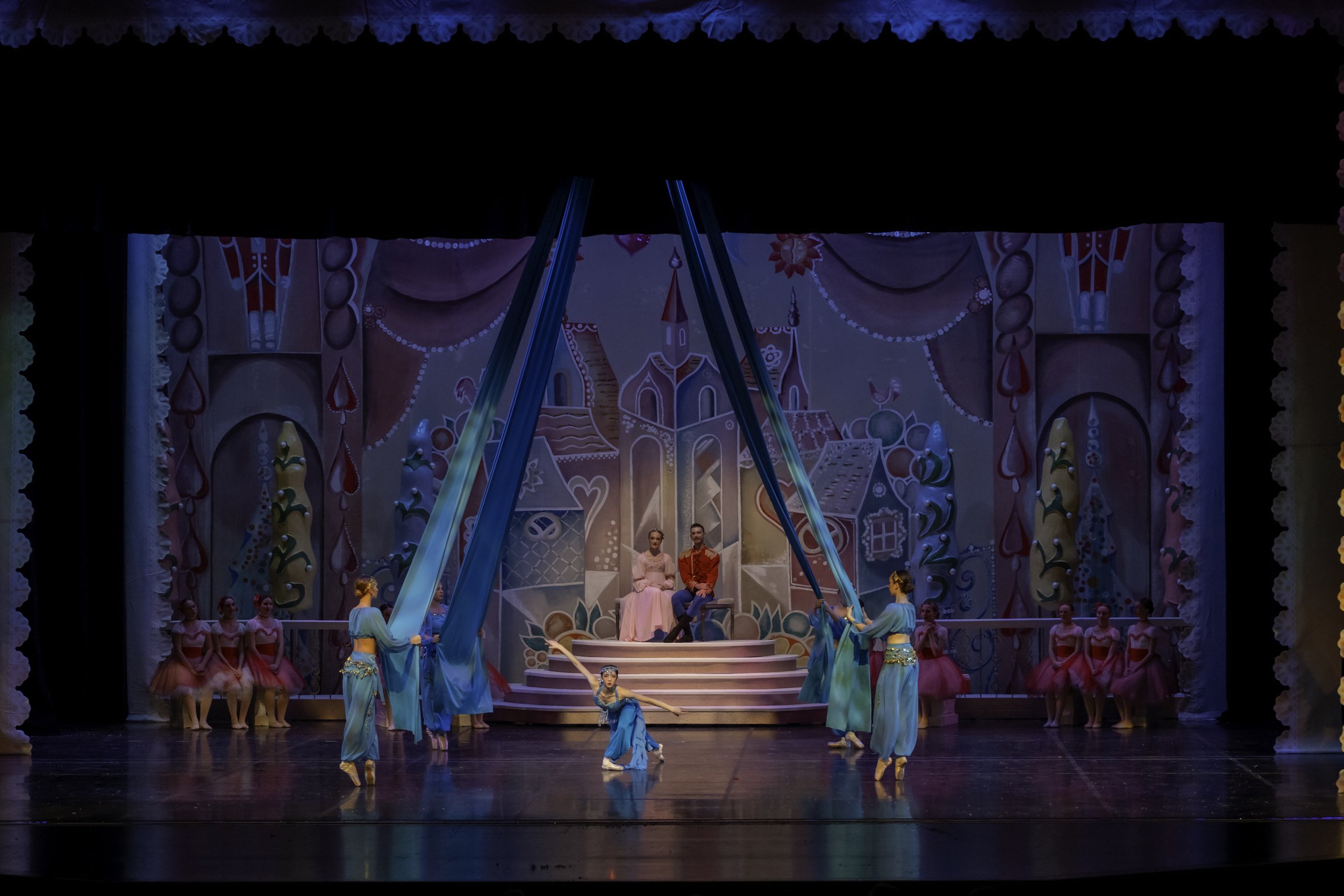 Ballet performance on stage with colorful backdrop, dancers in pink and blue costumes, some holding blue fabric long ribbons, while two dancers sit on elevated platform in center, surrounded by performers in pink tutus.