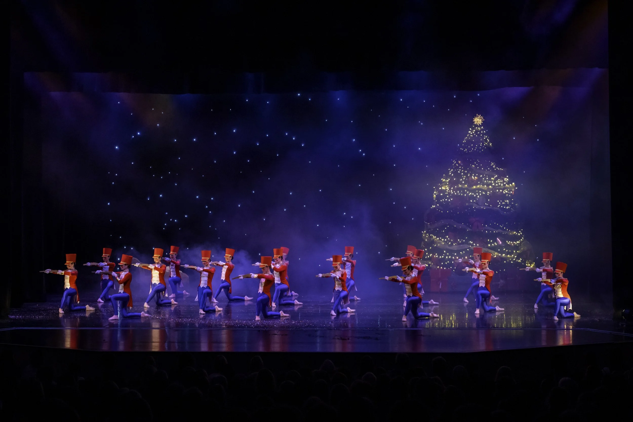 Children dressed as toy soldiers performing a Christmas-themed dance on stage with a decorated Christmas tree and starry sky backdrop.