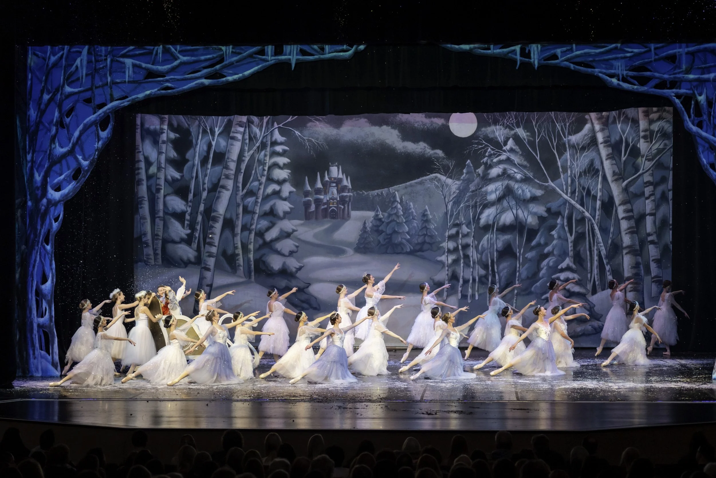 A ballet performance on stage with dancers in white tutus performing a dance in front of a wintery forest backdrop. The stage has blue tree props and a castle in the distance under a night sky with a full moon.