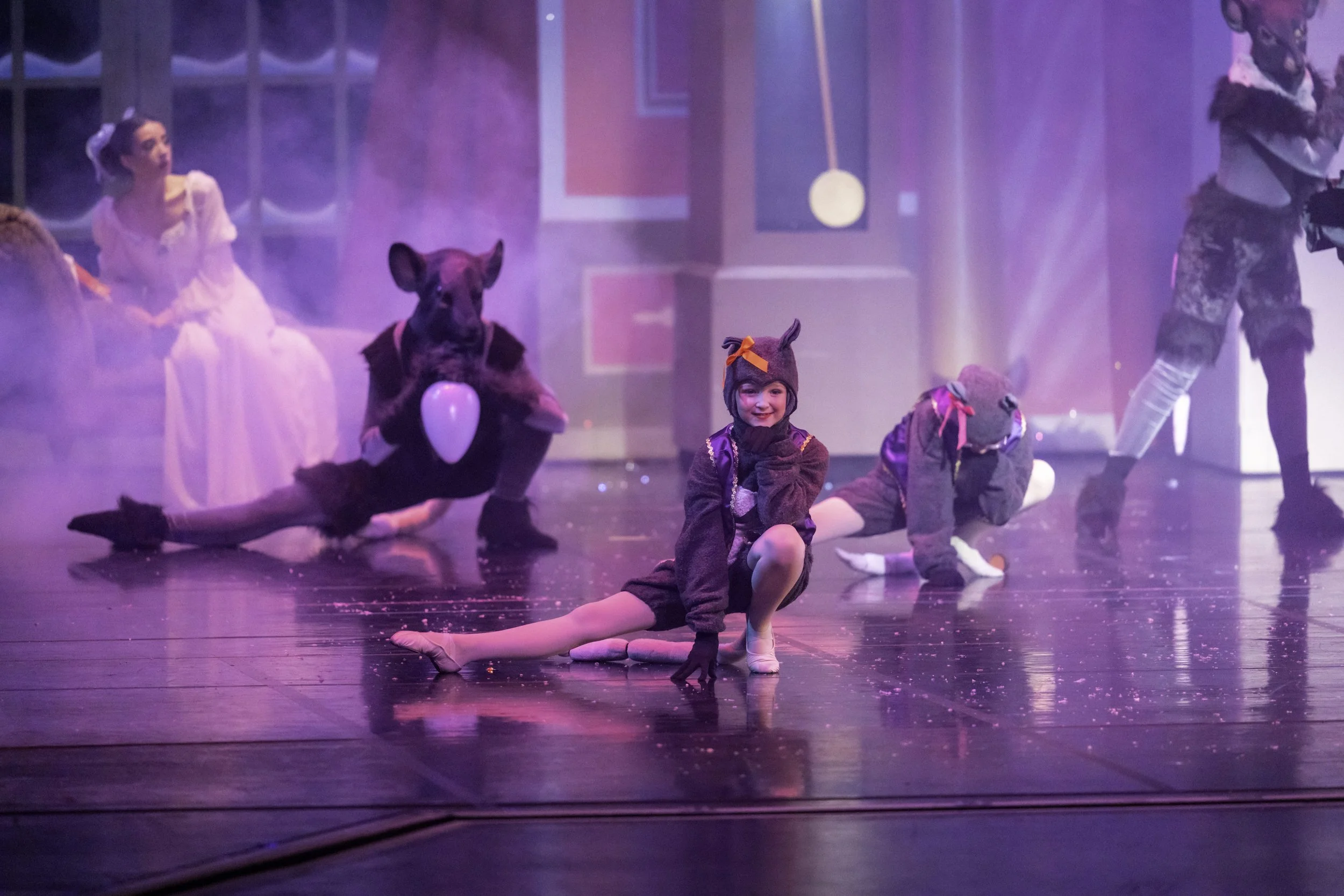 Children dressed in costumes perform on stage, with one girl in the foreground smiling and posing in a gray costume with a hat that has ears, and others dressed as a dog, a cat, and a woman in a light pink dress in the background, surrounded by purpl