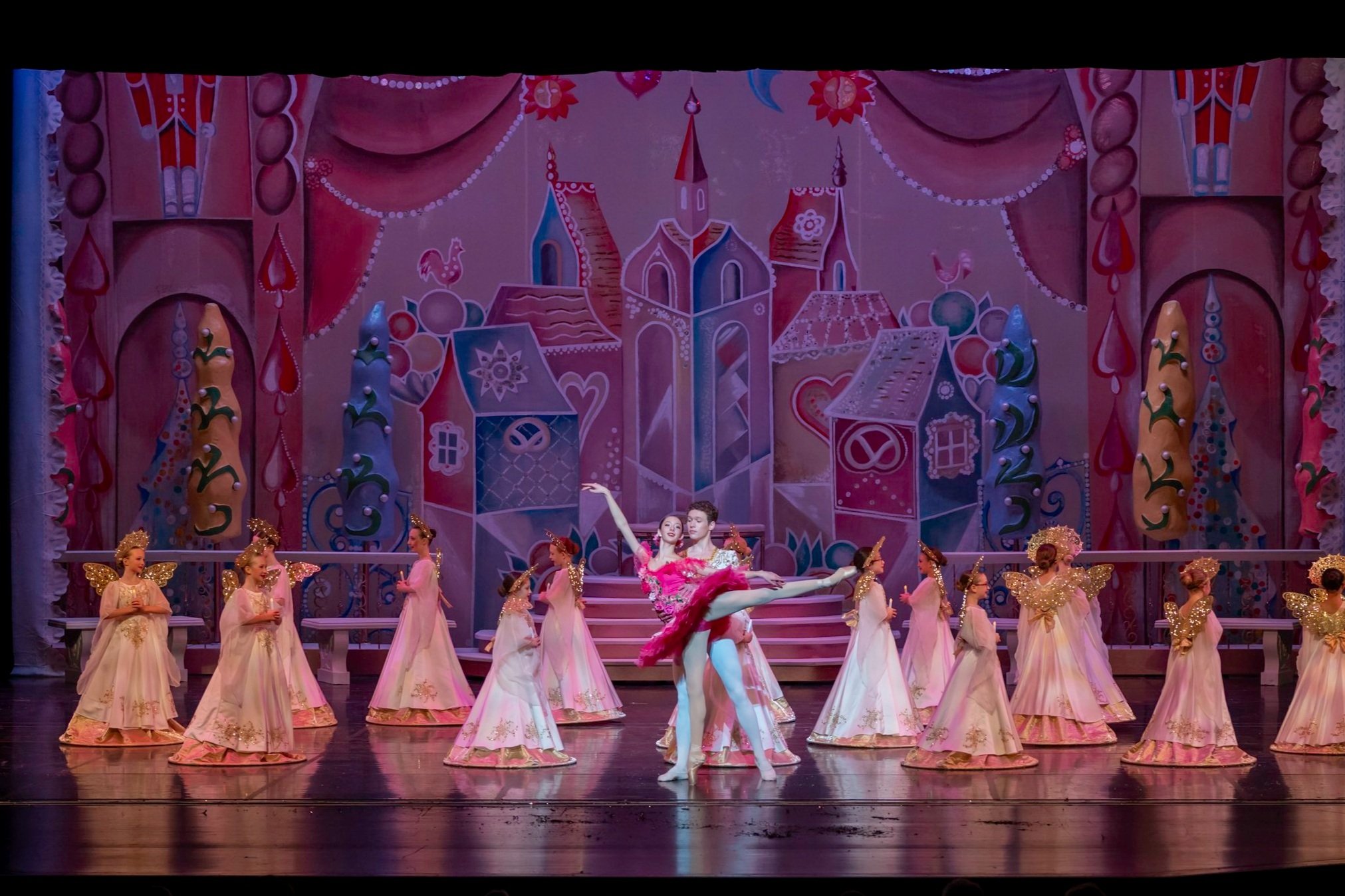 Ballet dancers performing on stage with a colorful, fantasy-themed backdrop featuring pastel-colored castles and trees, dressed in pink and white costumes with golden accents.