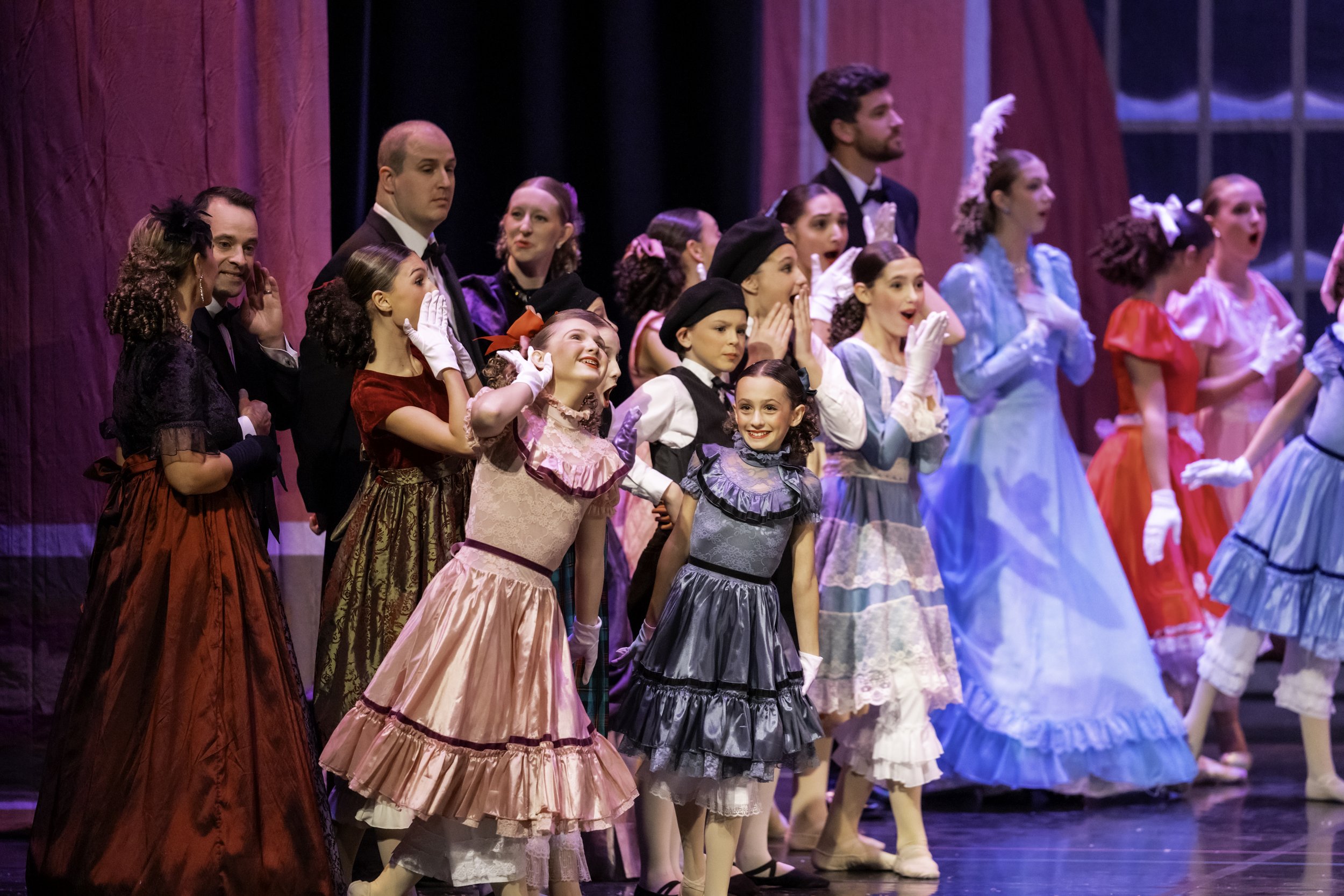 Cast of a musical or theatrical play dressed in colorful vintage costumes, standing on stage with purple curtains in the background.