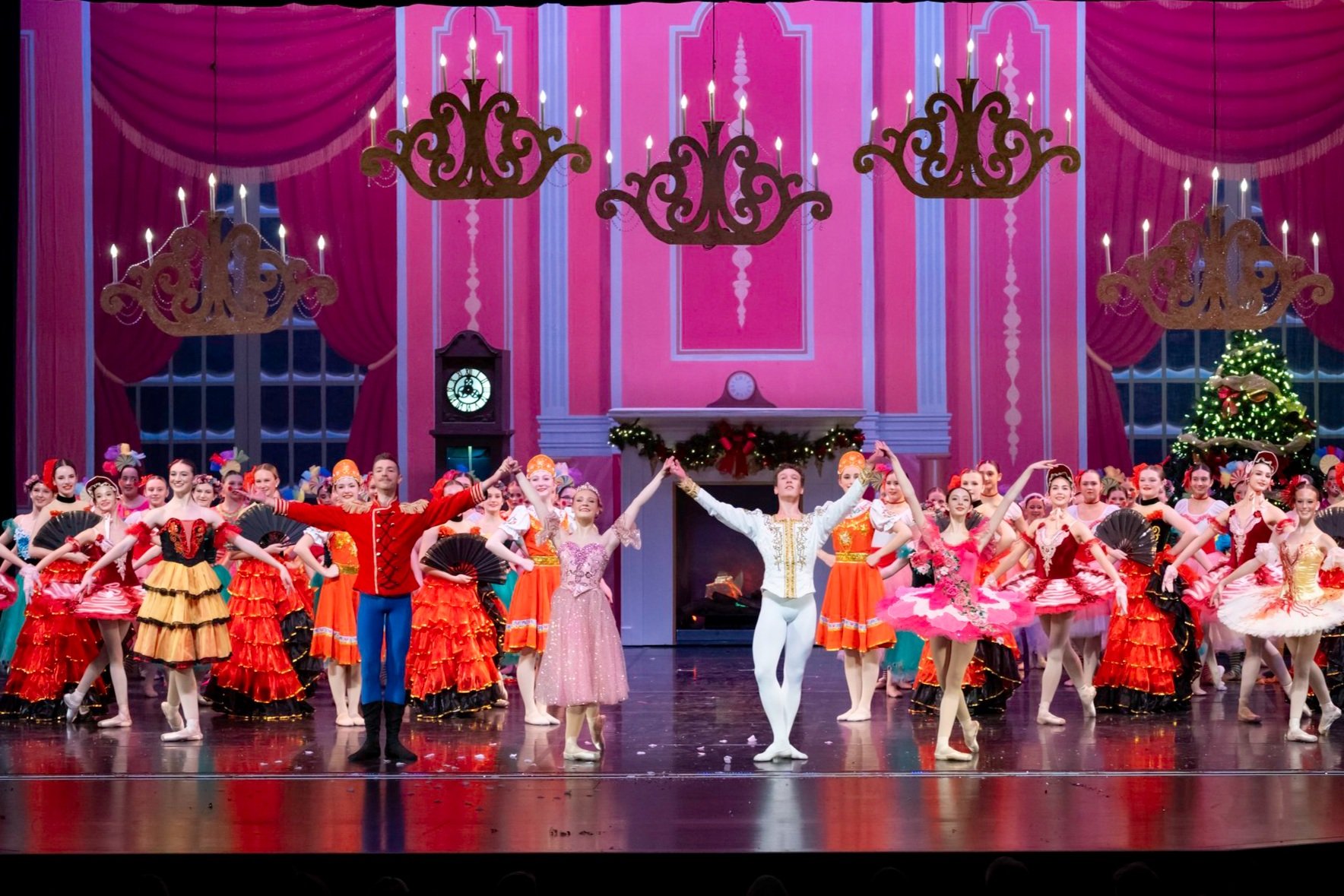 Ballet dancers on stage during a holiday performance, with festive decorations including a decorated Christmas tree and a fireplace surrounded by garland and ribbons.