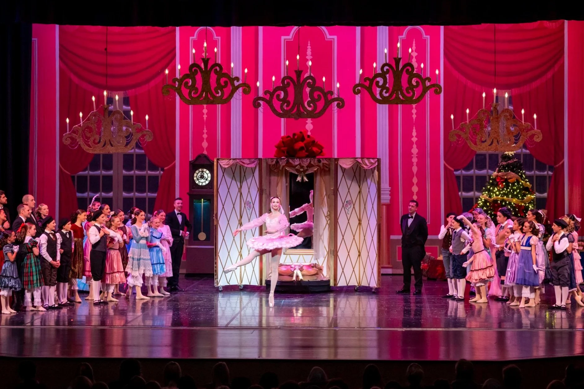 Stage scene from the Nutcracker ballet with dancers, children in colorful costumes, a Christmas tree, and ornate chandeliers in a festive setting.