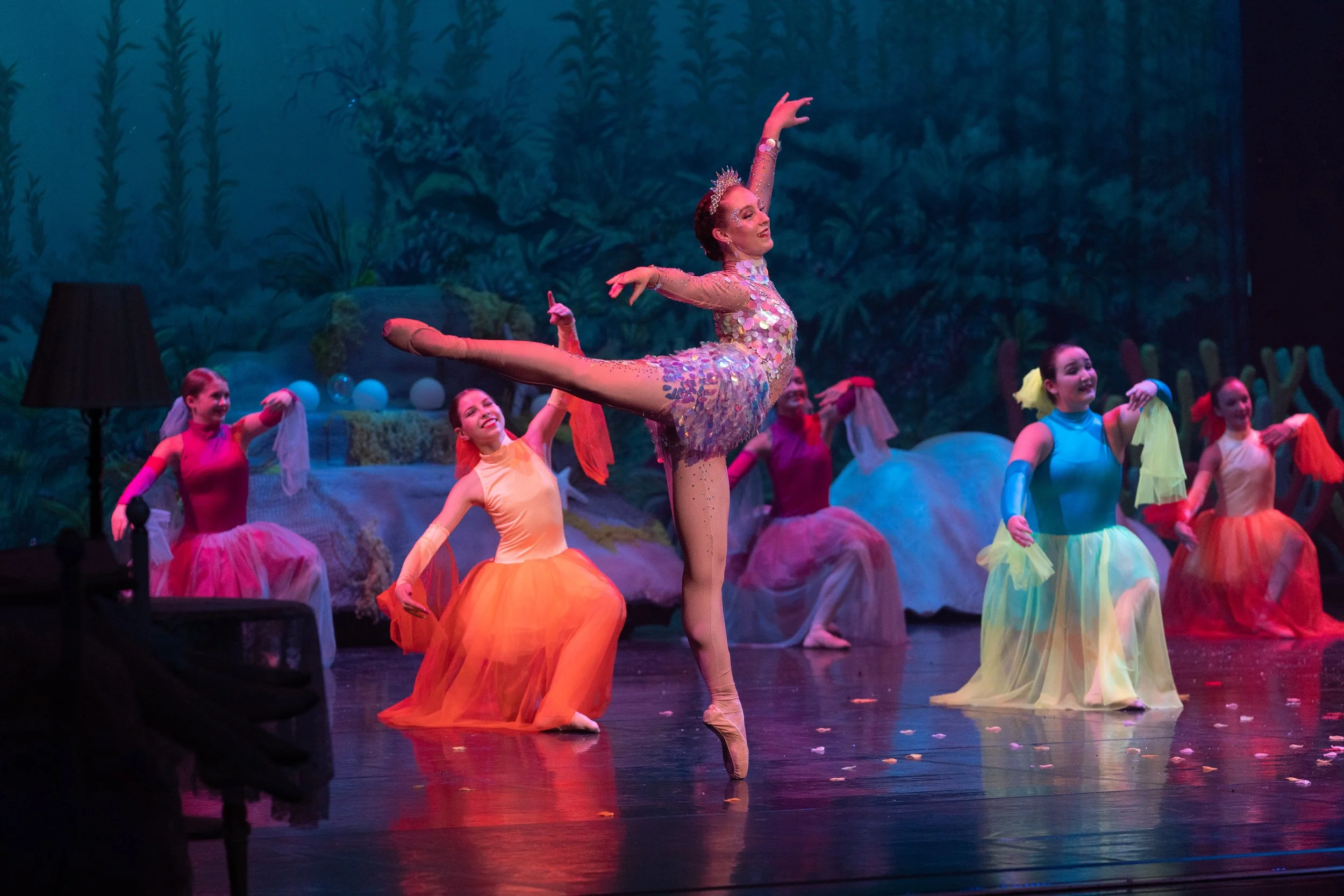 Ballet performance with a lead dancer in a glittery costume and colorful tutu, balancing on point with one leg extended, surrounded by fellow dancers in vibrant costumes on stage with a forest backdrop.