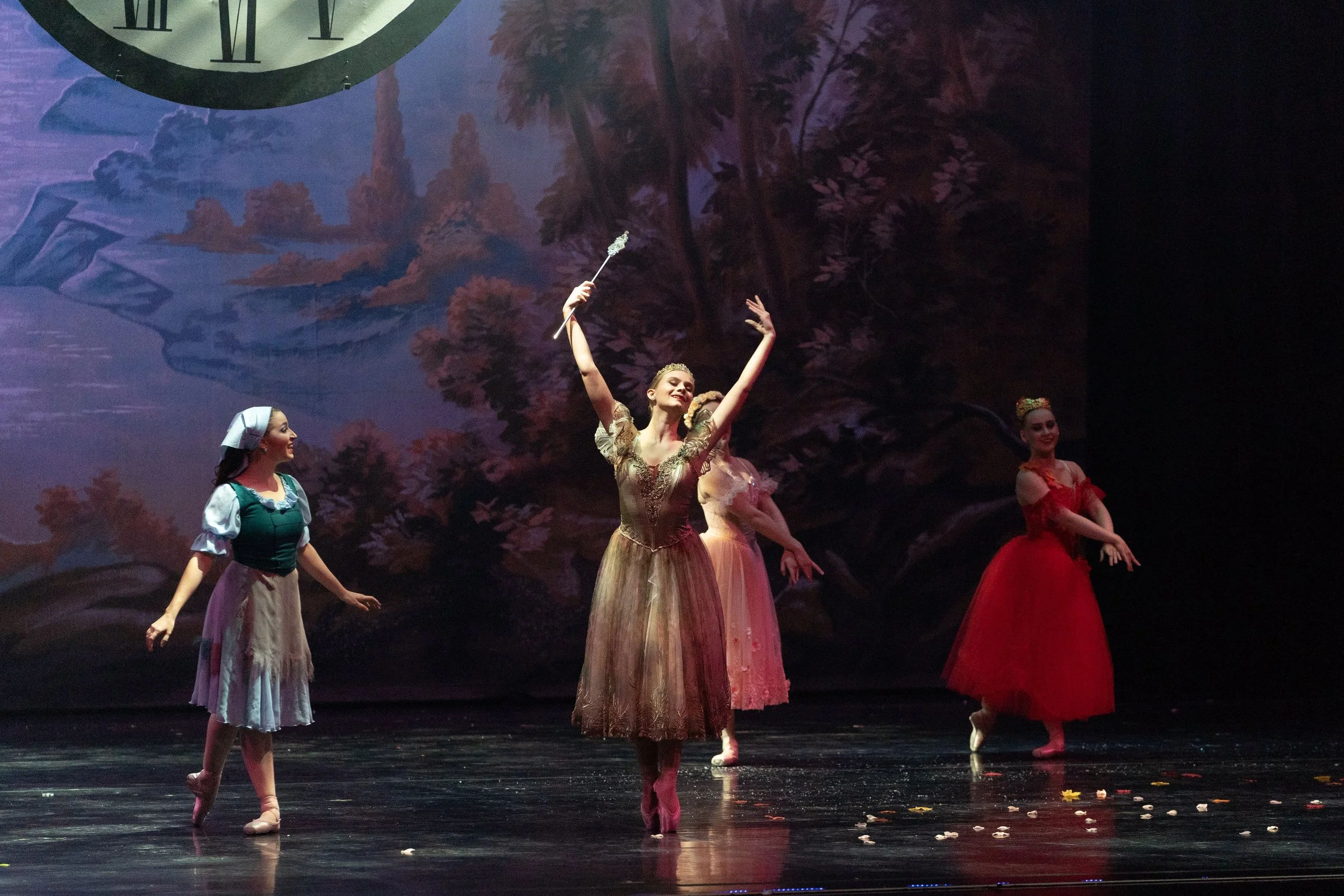 Four ballet dancers performing on stage with a scenic backdrop of a forest and river, one dancer in center holding a wand, others standing with arms extended at various angles.