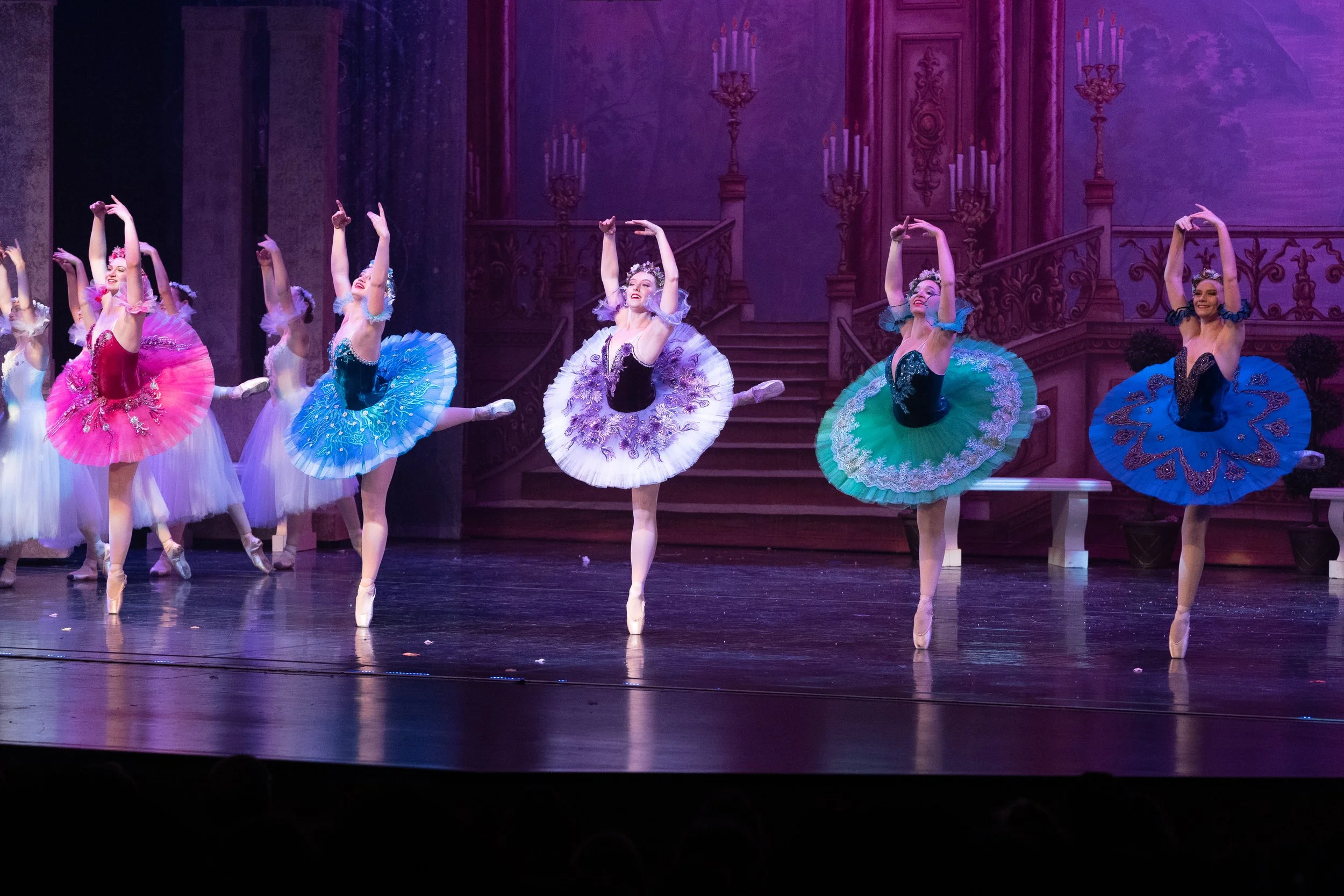 Ballet dancers performing on stage in colorful tutus, with a scenic background of stairs and ornate chandeliers.