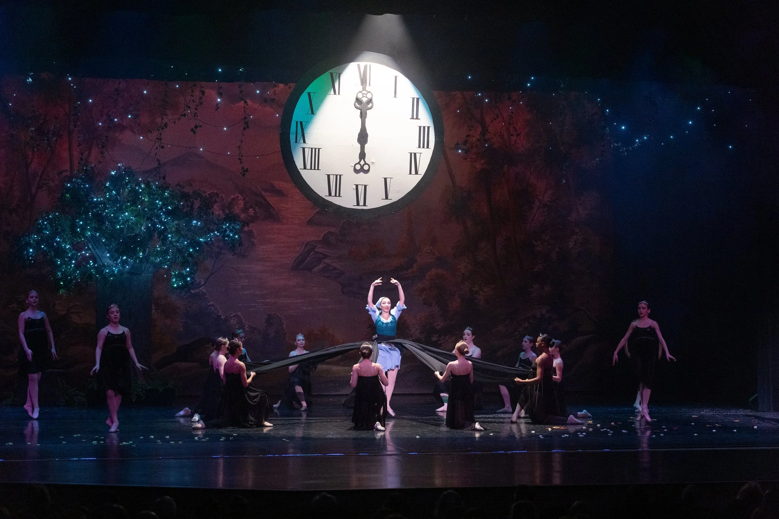 A ballet performance on stage with a large clock in the background showing midnight, surrounded by a painted scenic backdrop and illuminated tree. Dancers in black costumes perform, with one in a blue dress at center gesturing upward.
