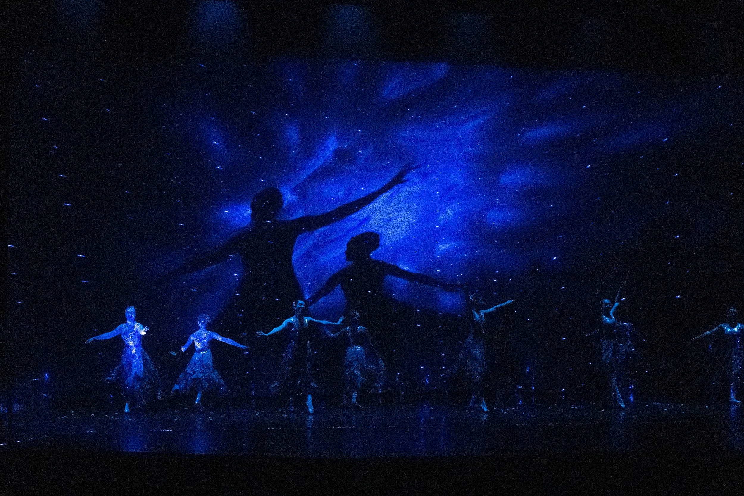 A dance performance with a backdrop of a starry night sky and two large silhouettes of dancers with arms outstretched, illuminated in blue light.