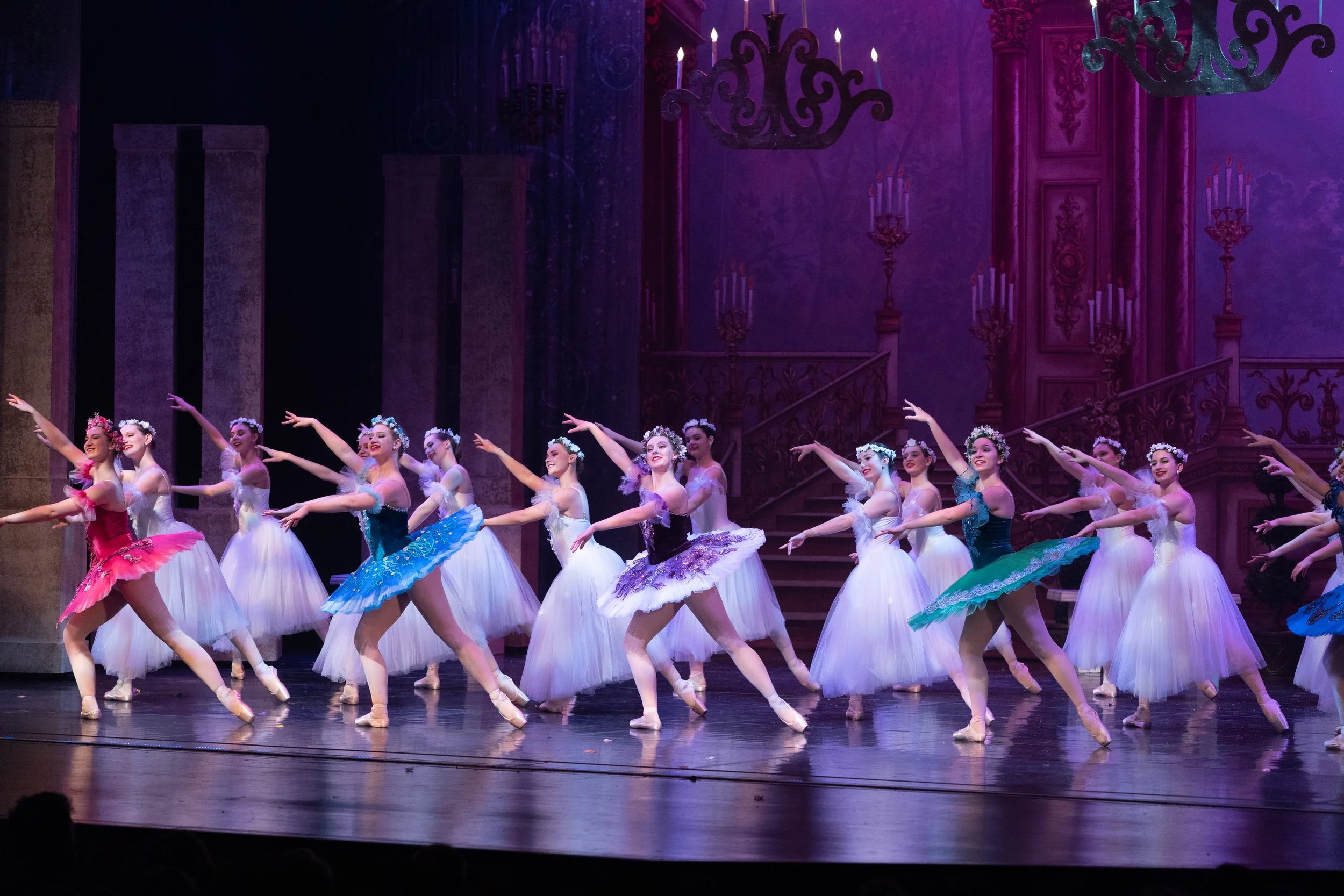 Ballet dancers performing on stage in colorful costumes with a purple and pink elaborate background.