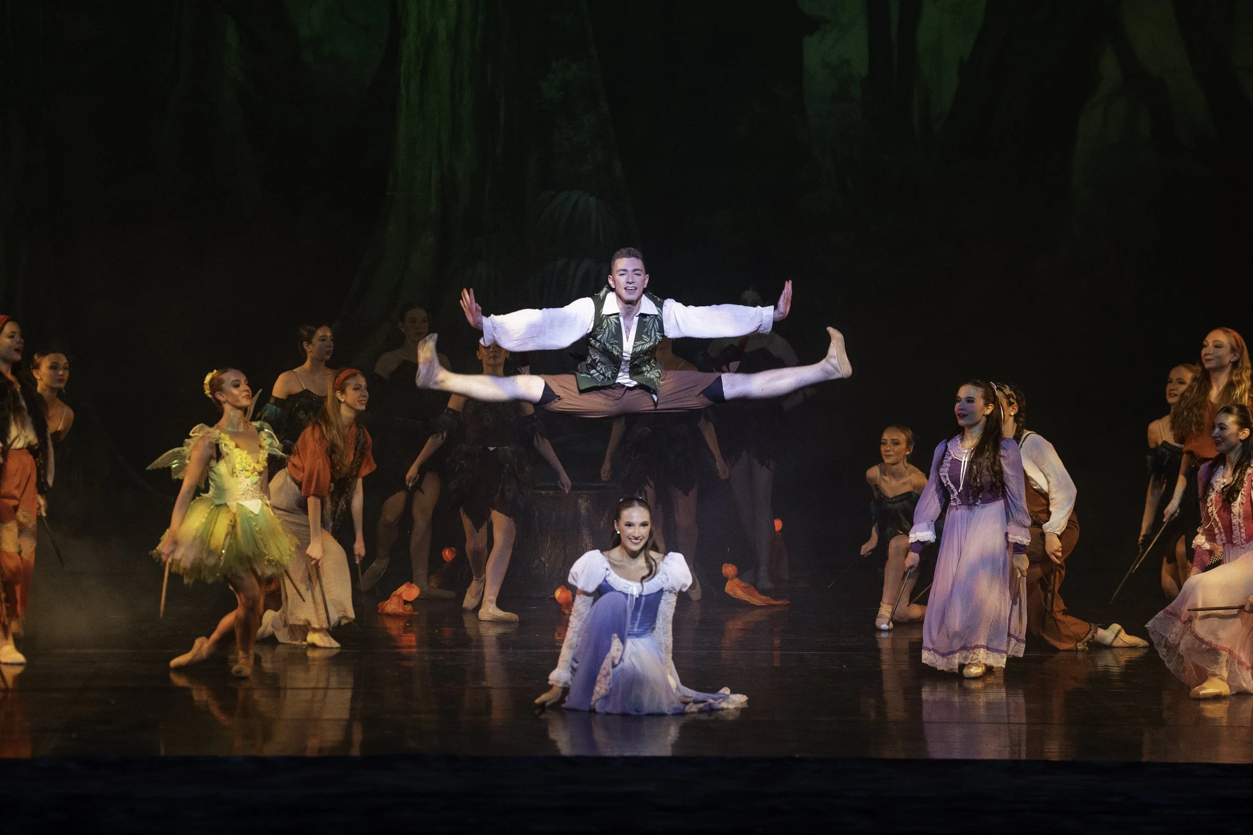 The image shows a stage performance with many dancers. They are dressed in colorful costumes, and one dancer is leaping above the others in a split position, with arms outstretched. The background includes a forest scene, and the stage is dimly lit w