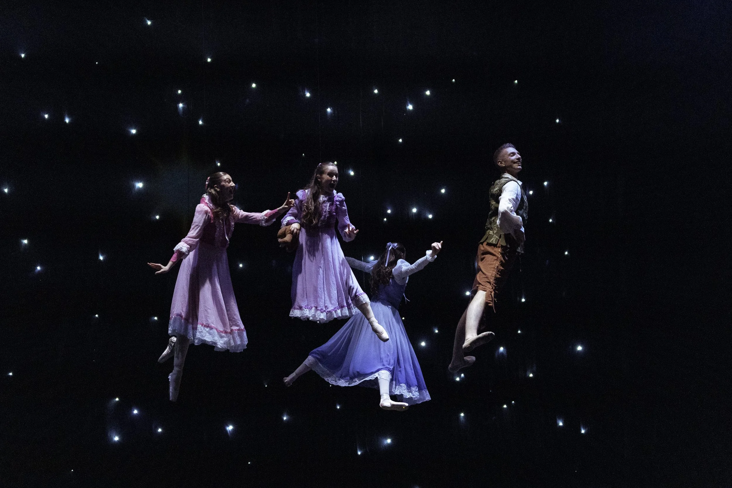 Four children in period costumes seemingly floating in a dark space with star-like lights, on stage during a theatrical performance.