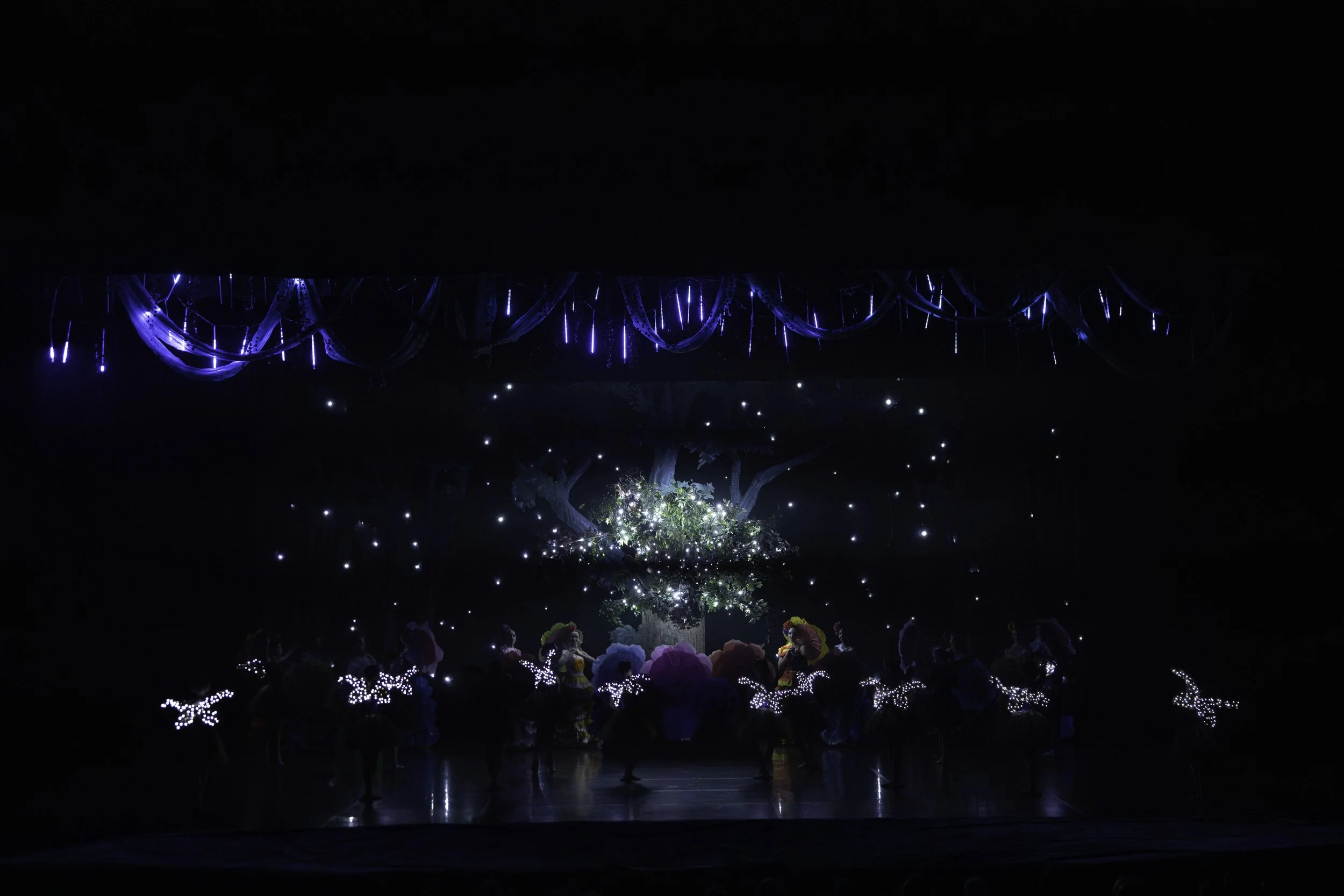 Stage scene with illuminated tree, dancers with glowing wings, and blue hanging decorations, creating a magical nighttime fantasy setting.