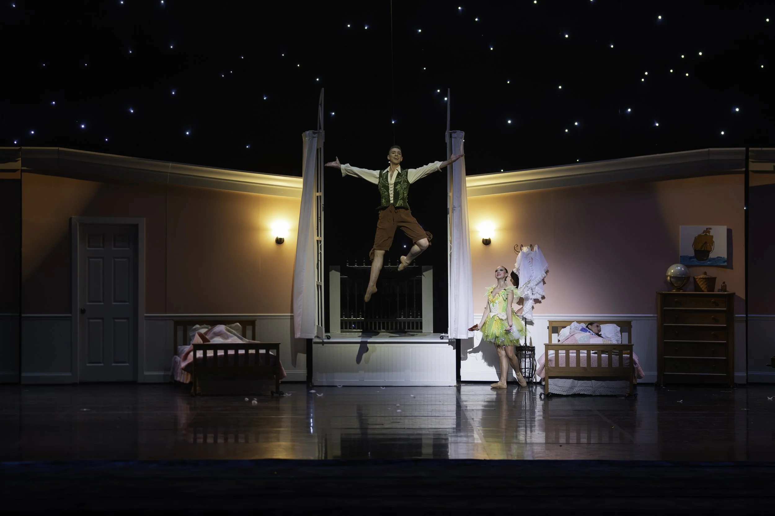 A stage scene from a theatrical performance set in a bedroom with pink walls, including beds, a dresser, and wall decor. A male performer is mid-air, hanging from a trapeze, with arms outstretched. Two female performers are on stage: one dressed in a