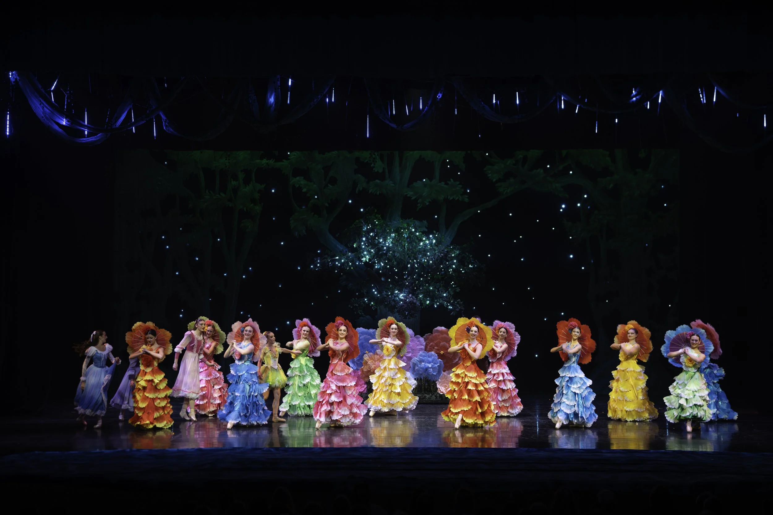 Colorful group of dancers wearing vibrant, floral-themed costumes performing on a stage with a backdrop of trees and a starry sky.