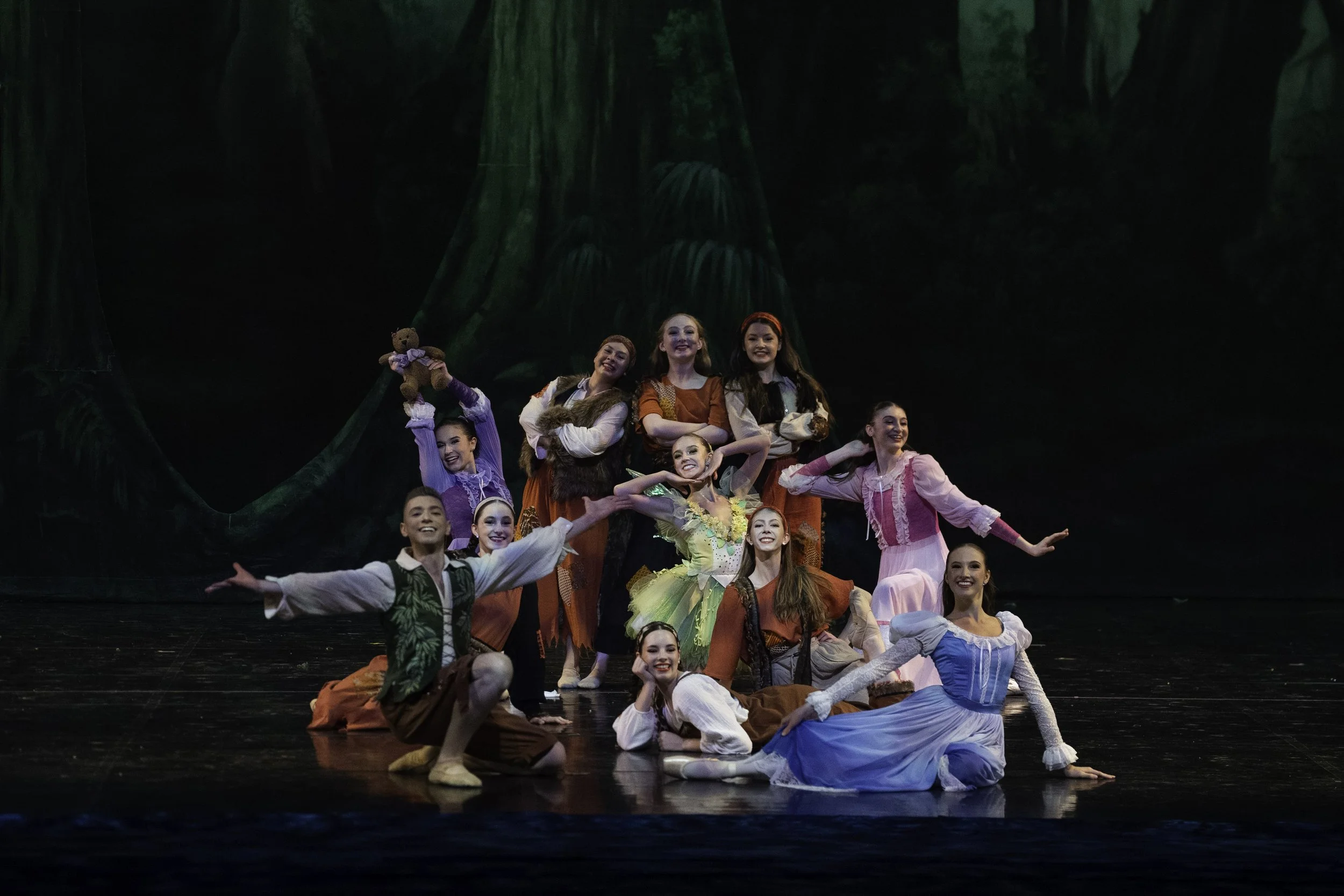 A group of young performers in colorful costumes on stage, posing for a cast photo during a dance or theater production, with a dark forest backdrop.