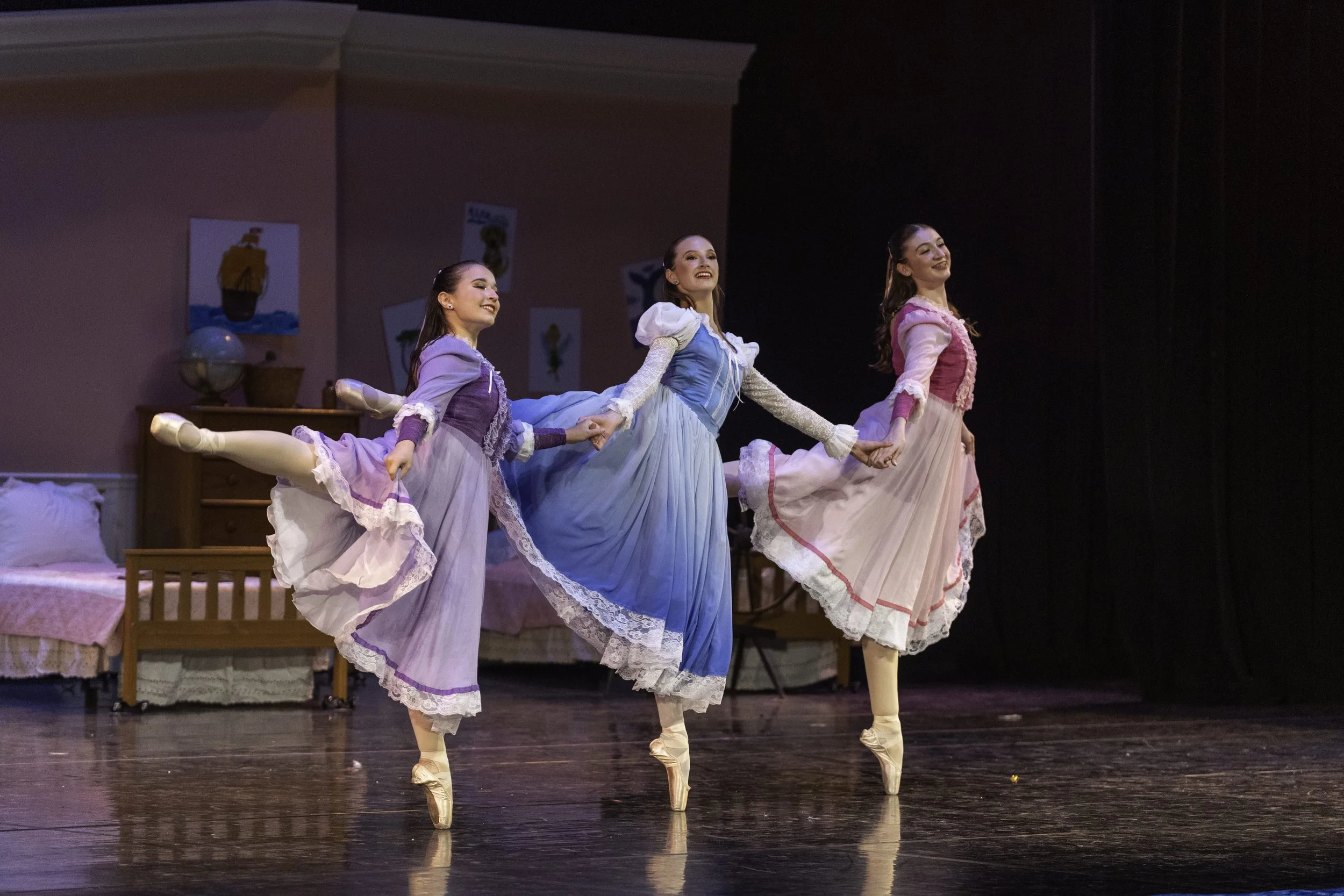Three ballerinas in pastel-colored dresses performing a dance on stage, holding hands and balancing on tiptoes, with a bedroom set and colorful artwork in the background.
