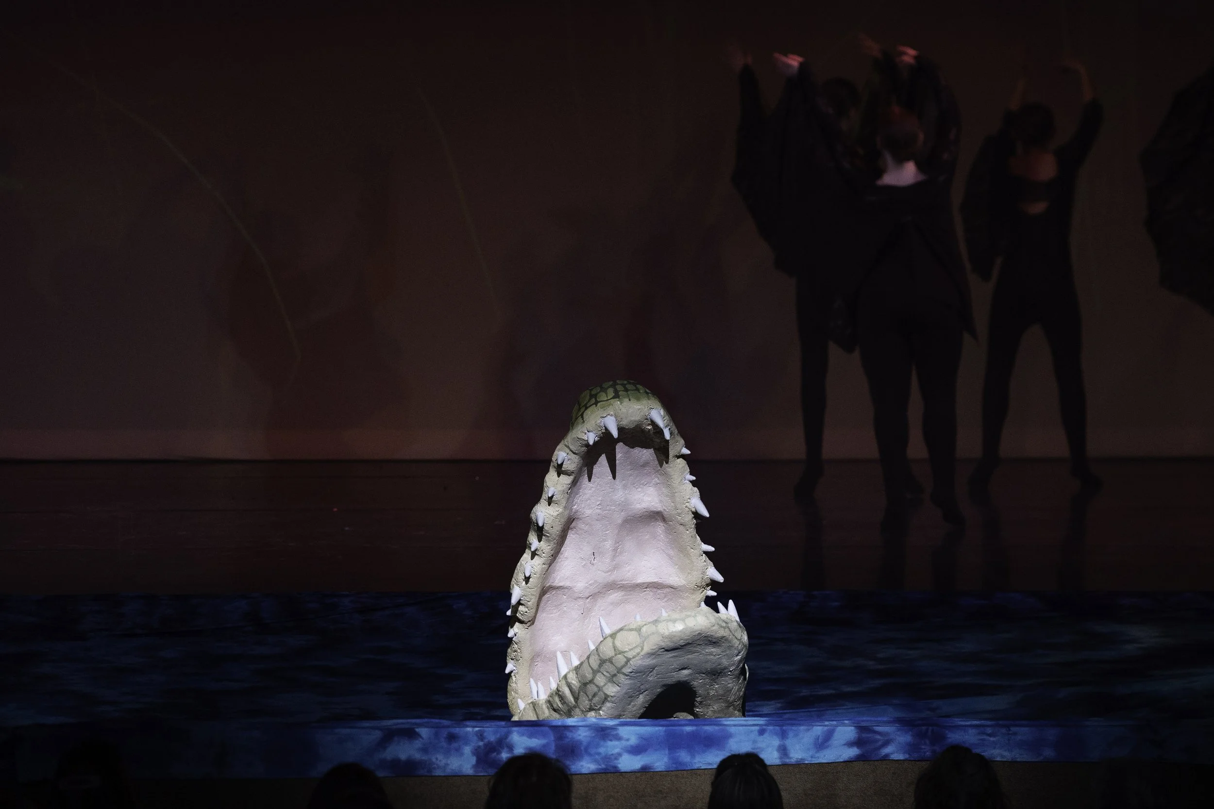 A stage with a crocodile prop with its mouth open, appearing to emerge from water, with dancers in black costumes in the background.