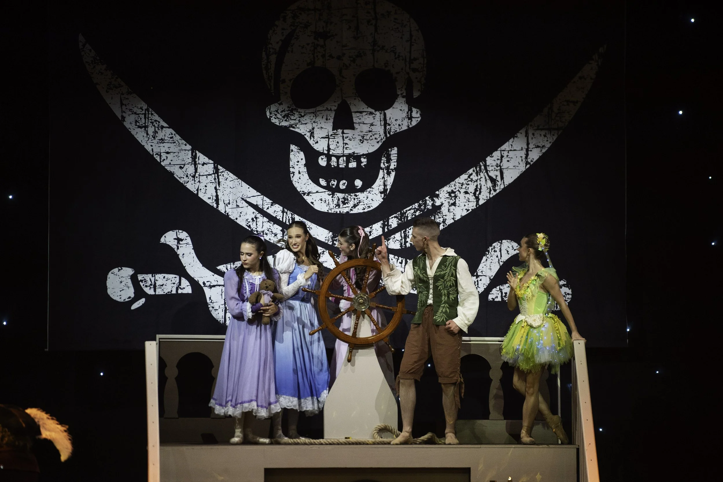 A theatrical performance of Peter Pan featuring five actors on stage, with a large skull and crossbones backdrop.