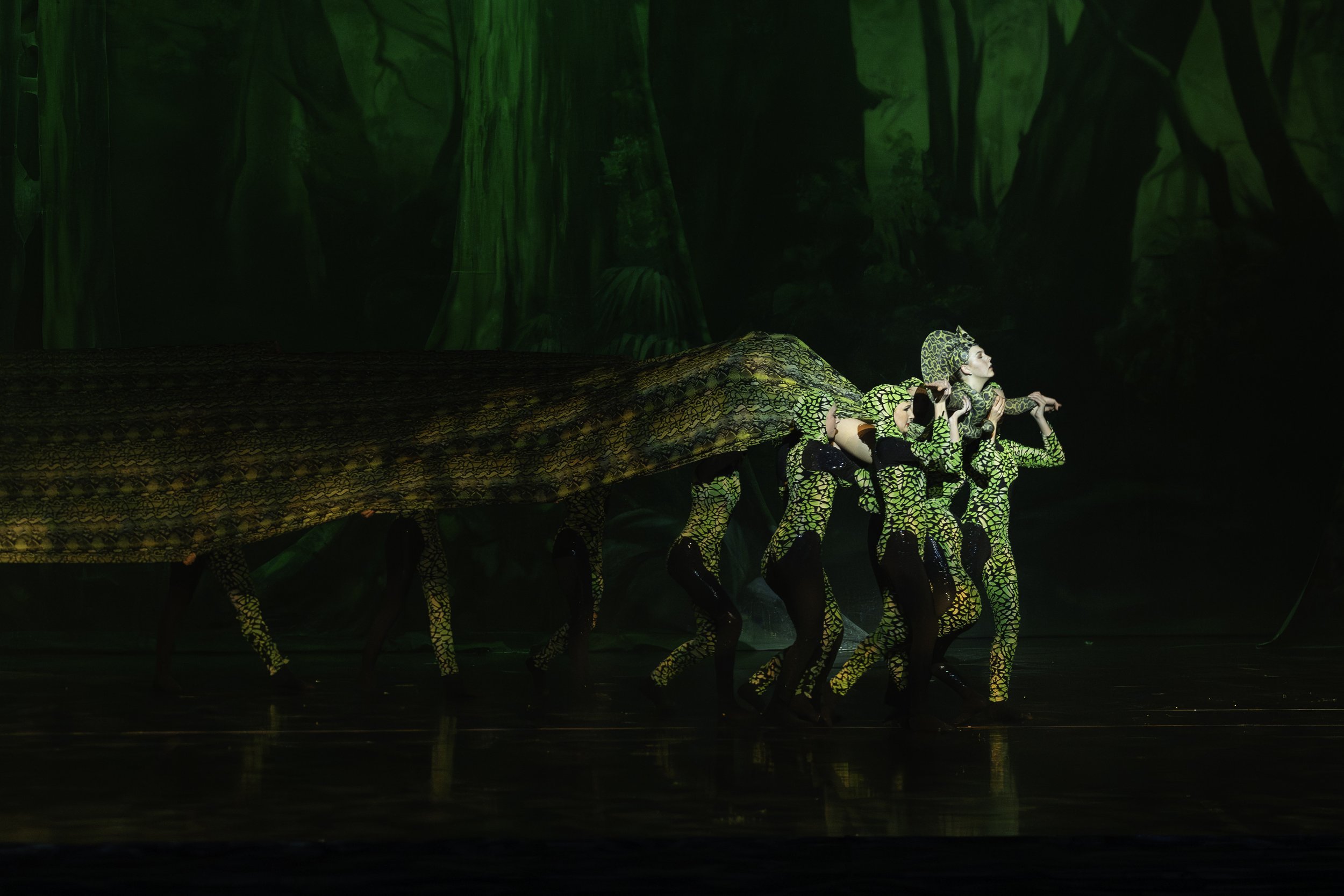 Performers dressed as snakes in a jungle-themed costume dance on stage with a backdrop of jungle scenery.