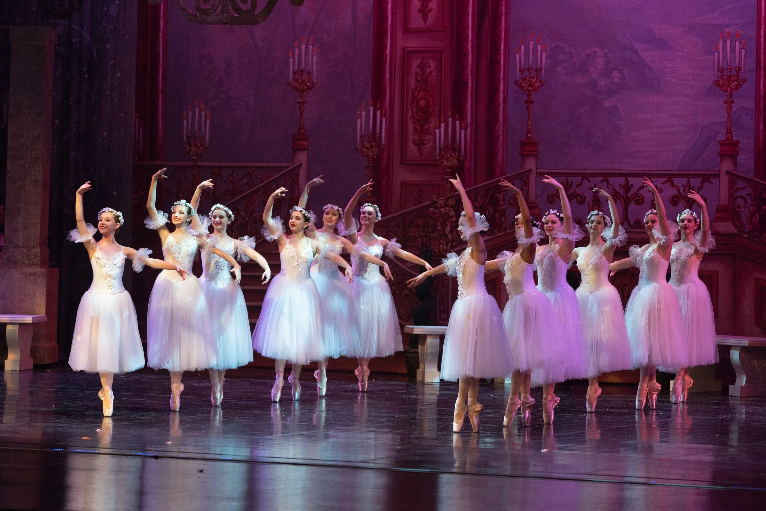 A group of ballerinas in white tutus performing on stage with pink lighting and ornate backdrop.