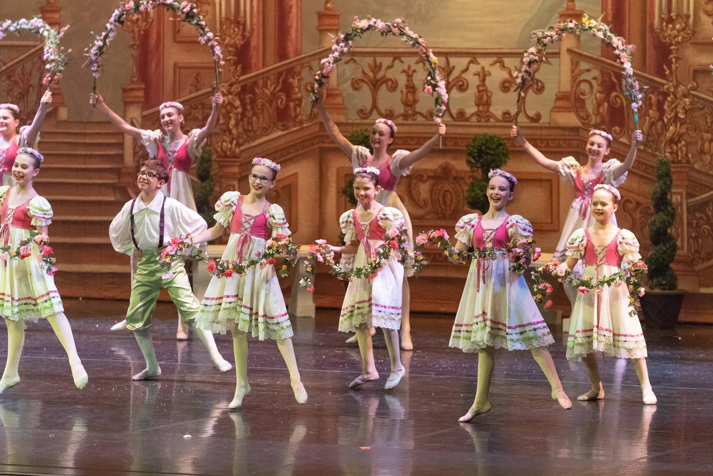 Children performing a ballet on stage, wearing pink and cream costumes with floral accents, with a decorated wooden staircase and floral arches in the background.