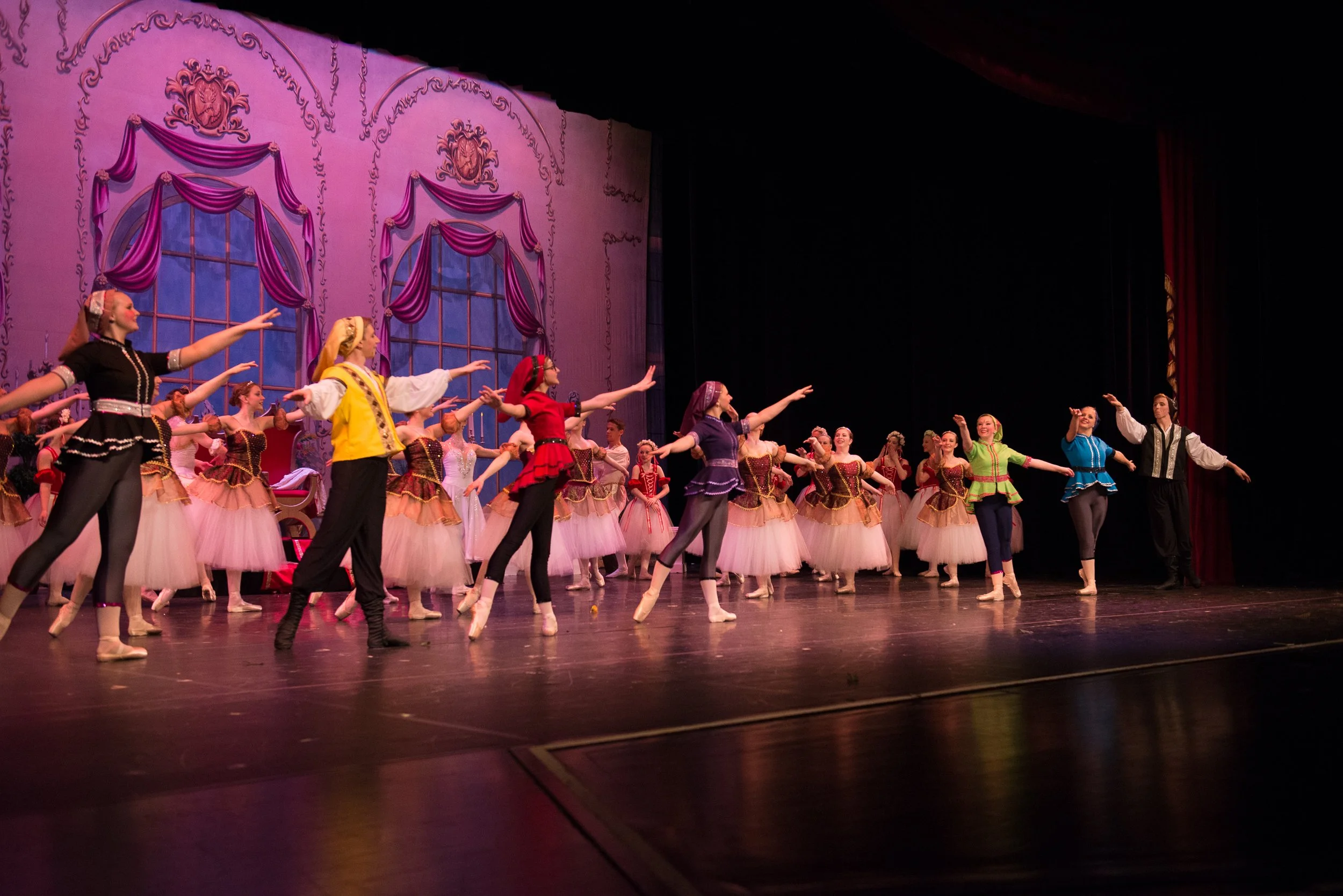 Ballet dancers perform on stage with pink and purple curtains and large windows in the background.