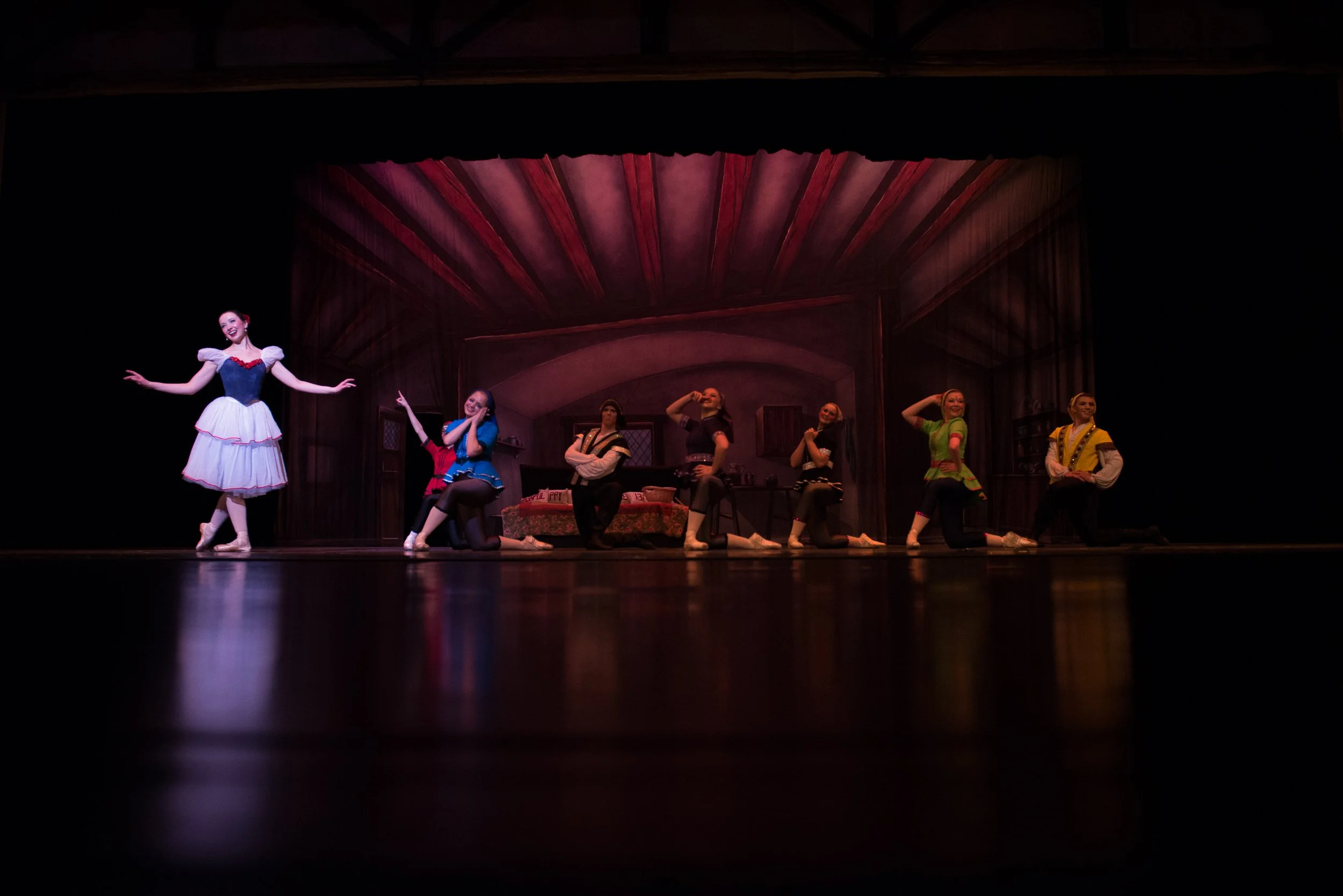 Theatrical stage scene with eight performers in colorful costumes, some kneeling and others standing, set against a detailed painted backdrop of a rustic interior. One performer stands apart, smiling with arms outstretched.
