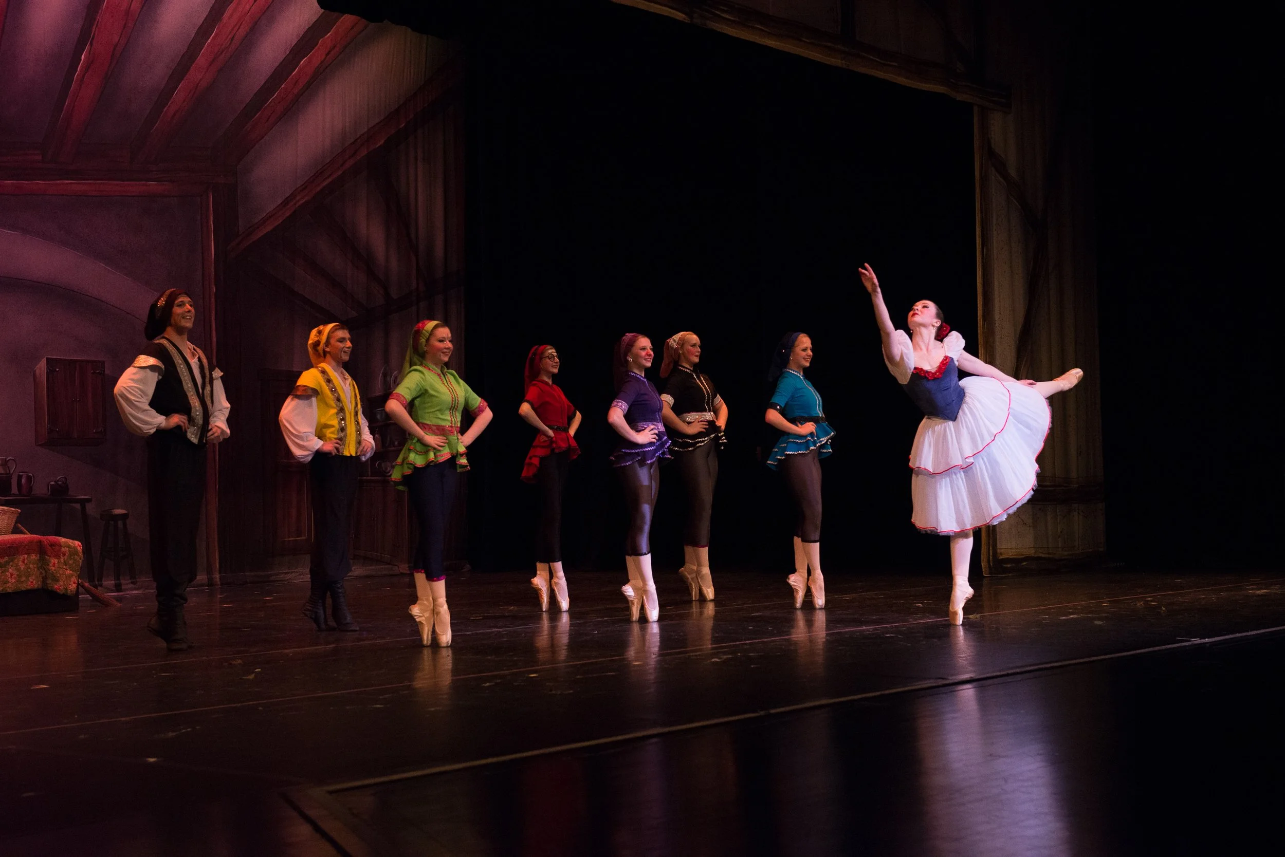 Ballet dancers performing on stage with one dancer in a white tutu and black vest executing a pointe move, while seven other dancers in colorful costumes observe.