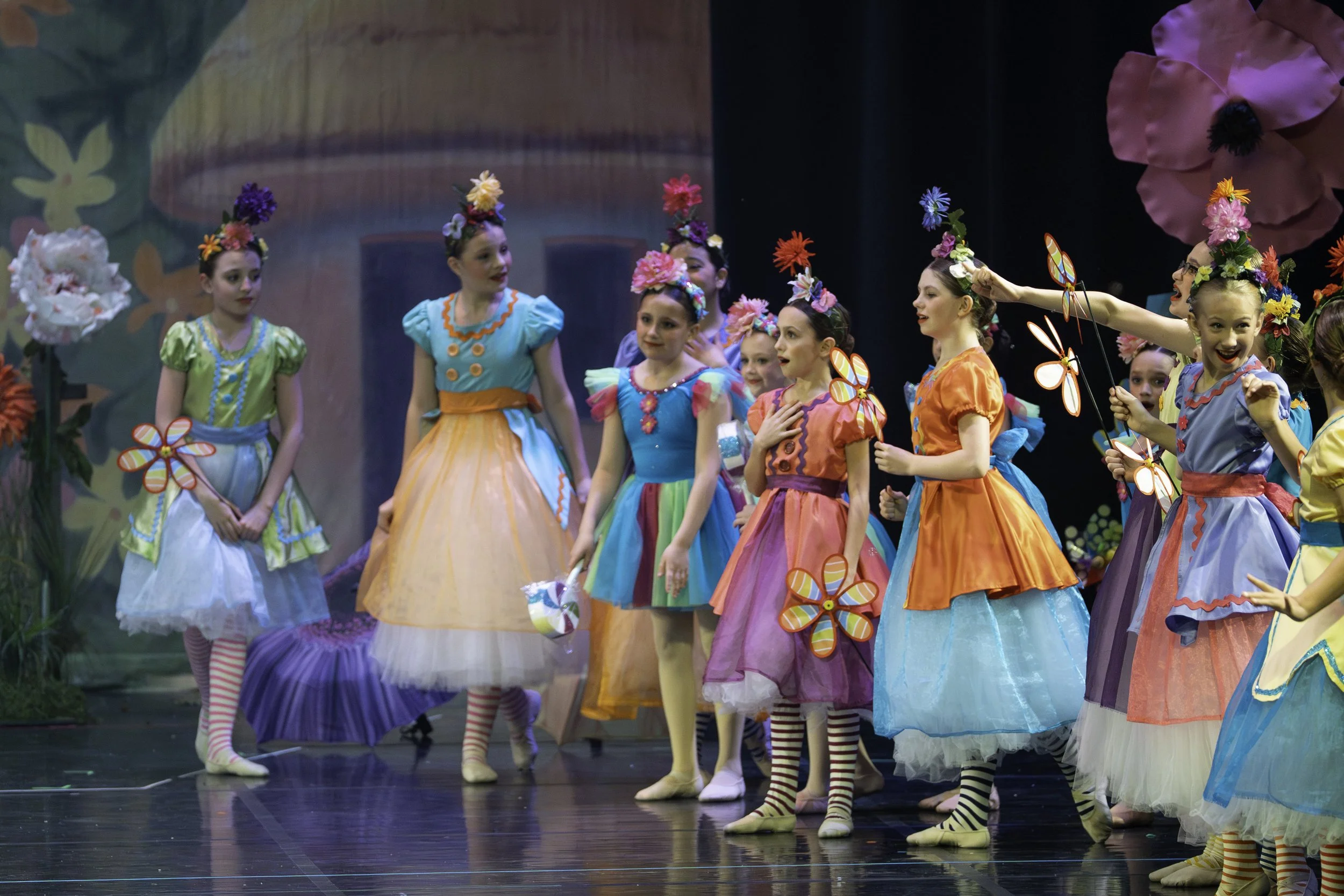 Young girls dressed in colorful costumes and flower headpieces performing on stage with a painted backdrop.