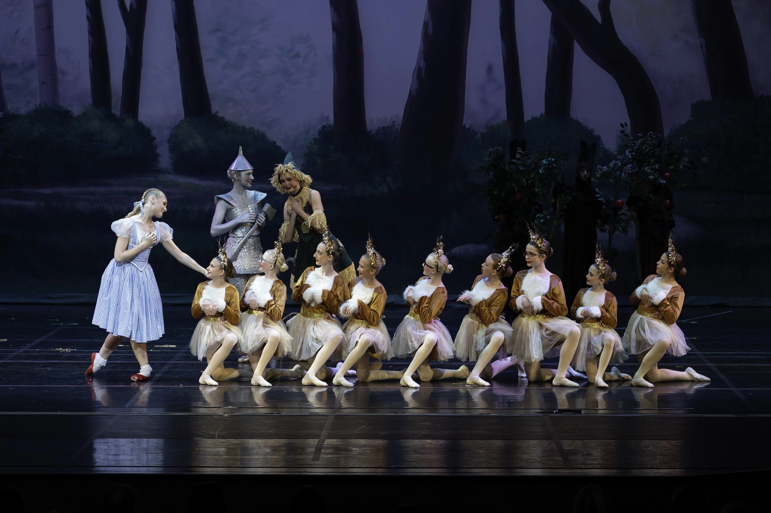 Children dressed as mice performing a ballet on stage, with characters from The Wizard of Oz, including the Tin Man and the Wizard, in a forest-themed backdrop.