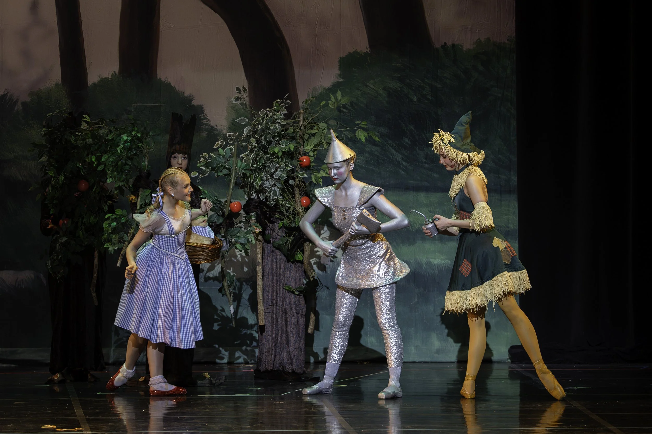 Children performing a theatrical play with characters dressed as characters from The Wizard of Oz, including Dorothy, the Tin Man, and the Scarecrow, on stage with a painted forest background.