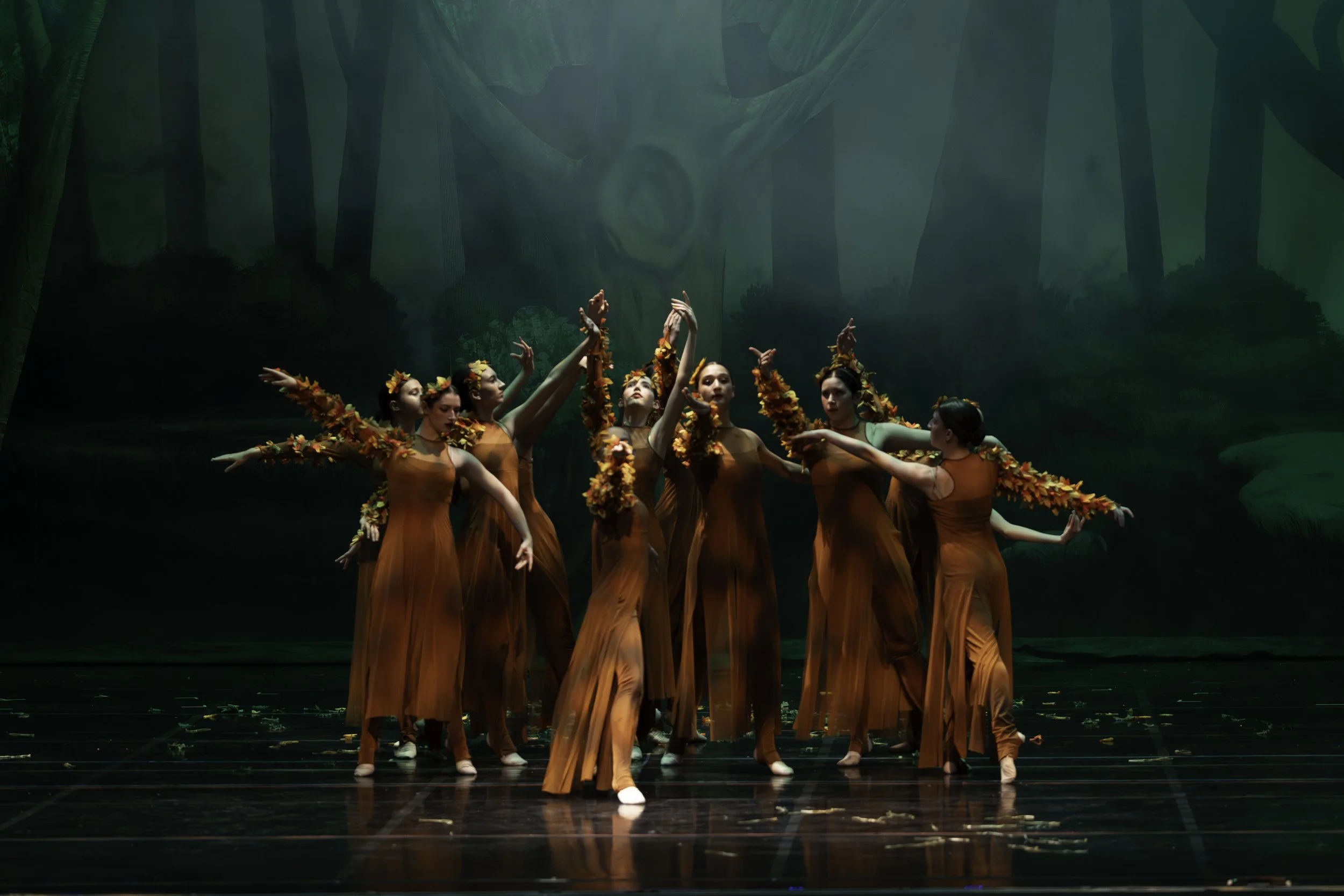 Group of dancers dressed in orange costumes with fall leaves, performing on a stage with a forest backdrop.