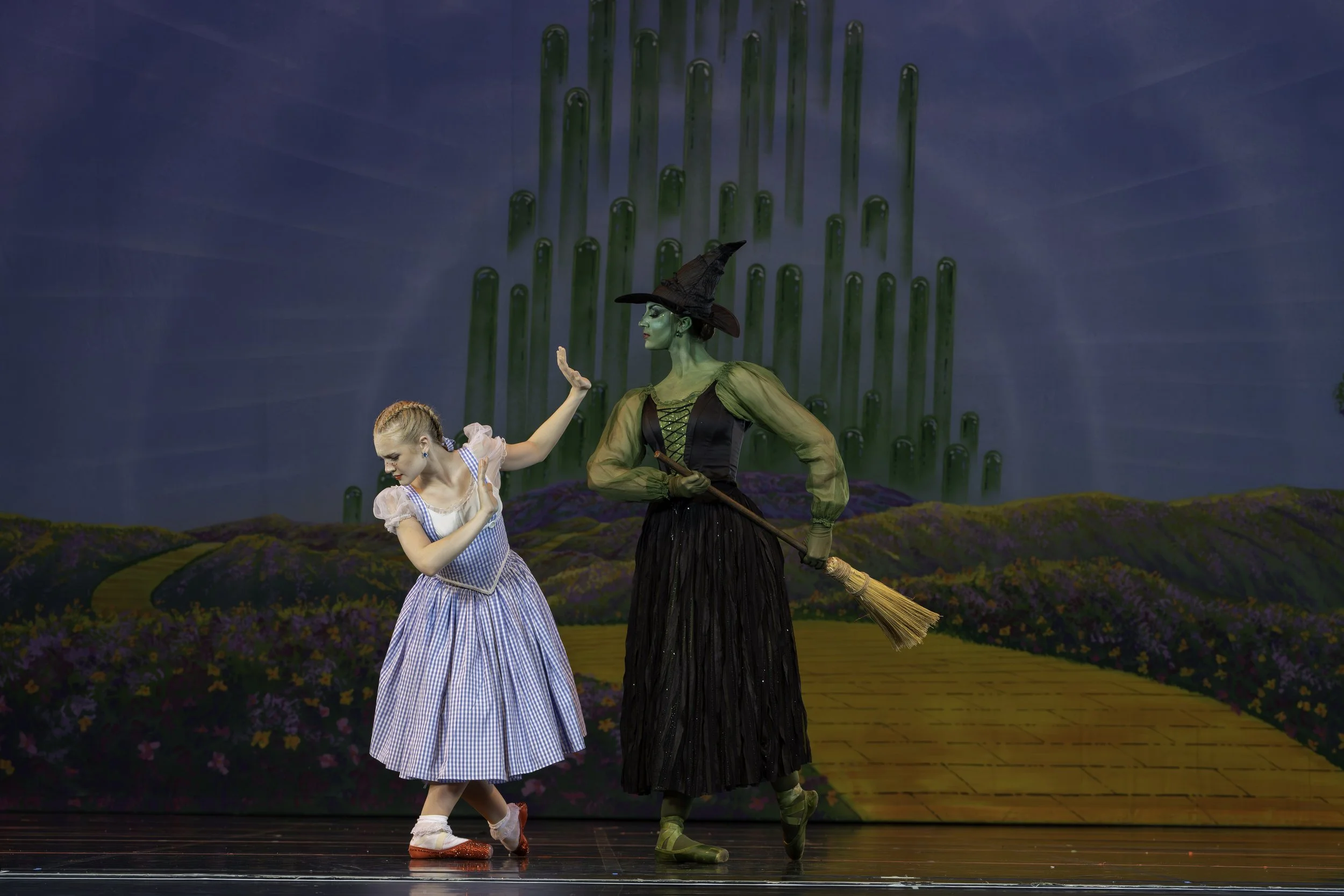 A girl dressed as Dorothy from The Wizard of Oz dancing with the Wicked Witch of the West, holding a broomstick, on a stage with a colorful painted backdrop of a yellow brick road and green tornado.