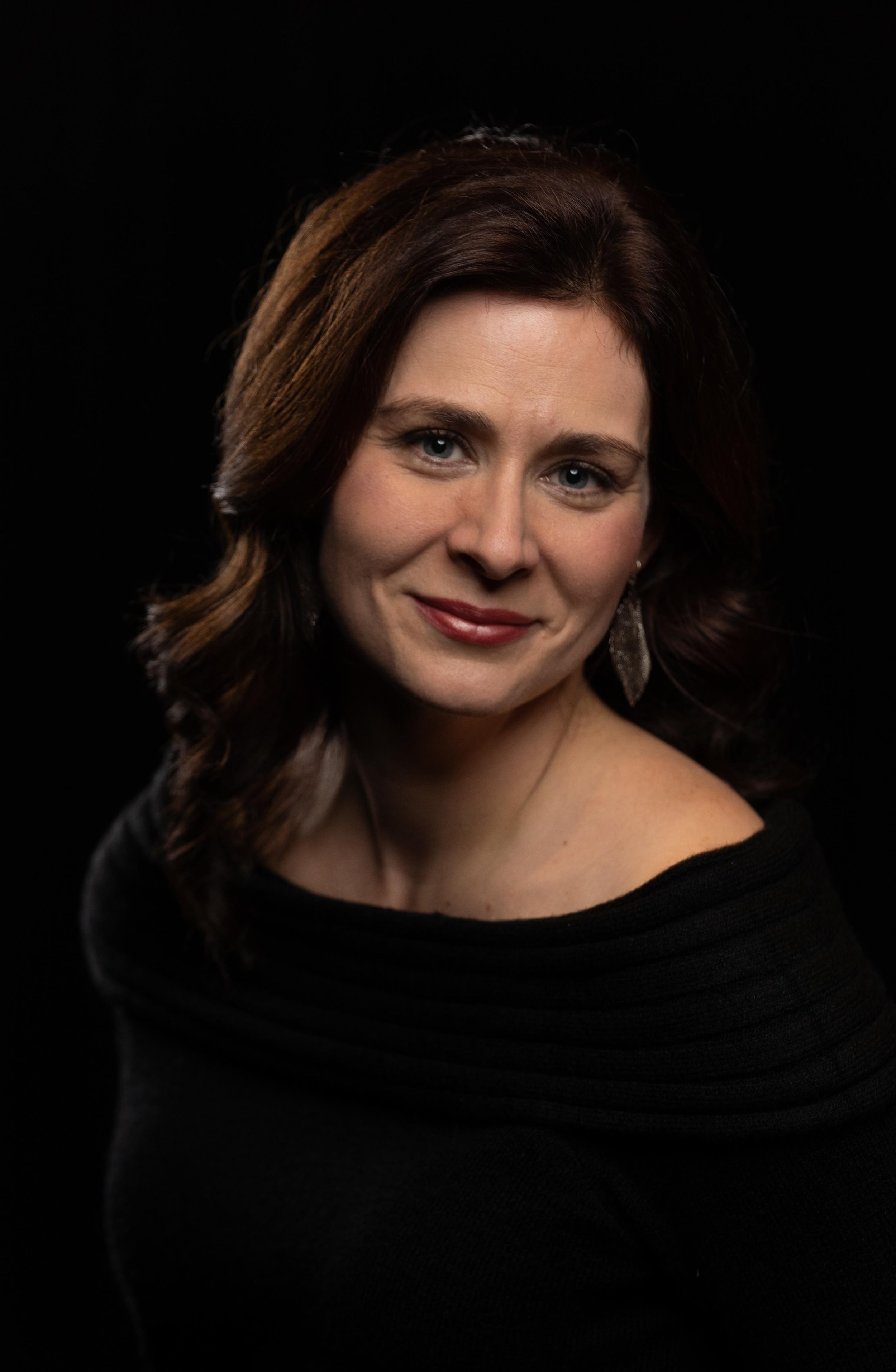 A woman with chestnut brown hair and blue eyes wearing a black off-the-shoulder top and silver earrings, smiling softly against a black background.
