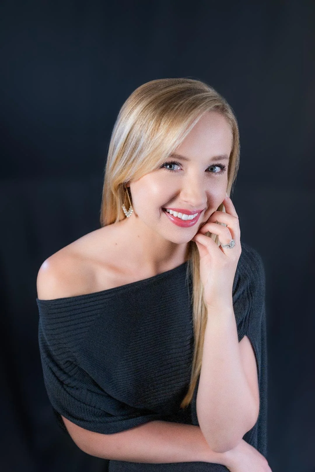 A young woman with blonde hair and blue eyes smiling, wearing a black off-the-shoulder top, earrings, and a ring, against a dark background.
