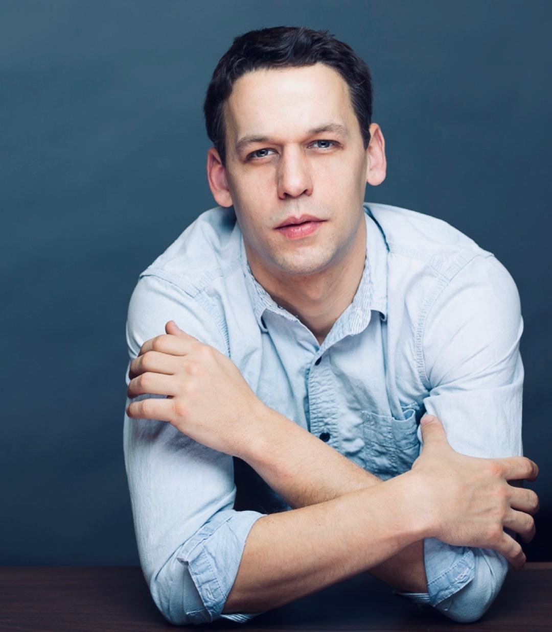 A young man with dark hair and blue eyes, wearing a light blue button-up shirt, poses with his arms crossed over each other against a dark background.