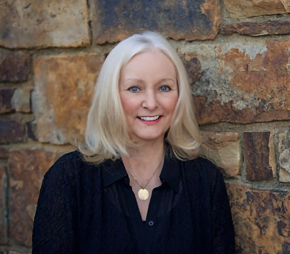 A woman with shoulder-length blonde hair, blue eyes, and a friendly smile, wearing a black textured blouse and a gold necklace with a round pendant, standing in front of a rustic stone wall.
