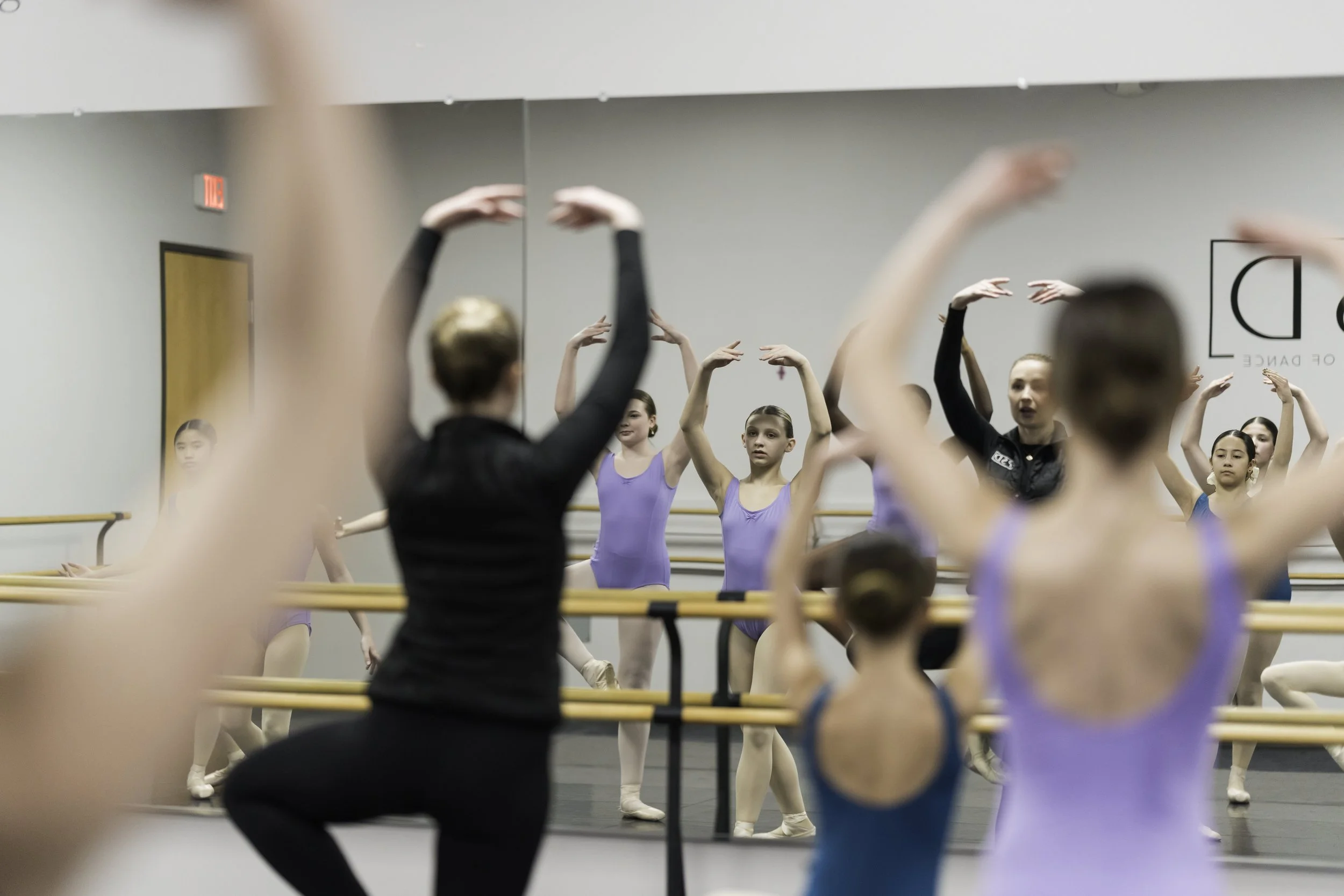 Ballet students practicing at a dance studio, guided by an instructor, with a mirror reflecting their movements.