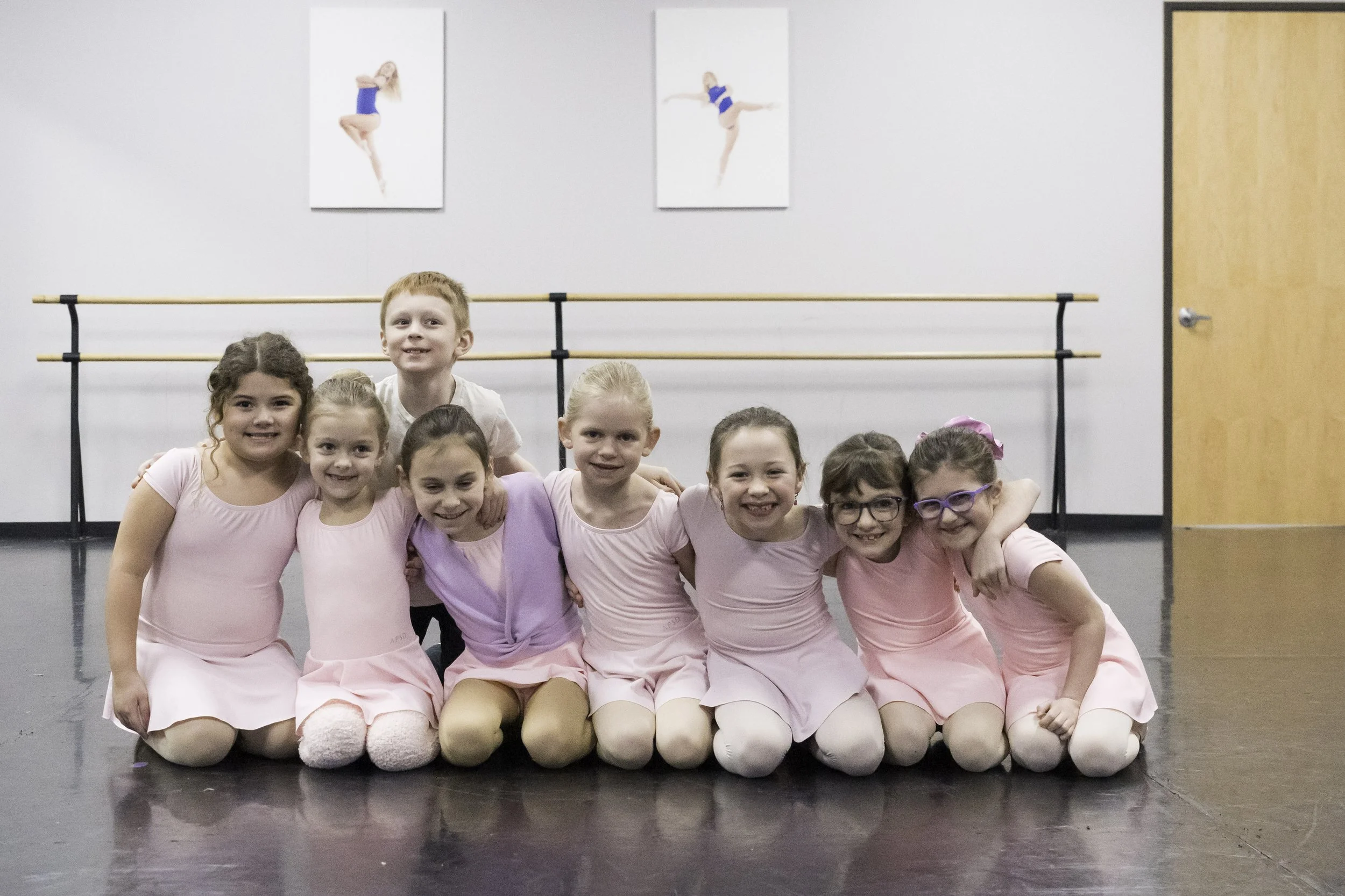Group of young girls in pink costumes at ballet studio posing with arms around each other.