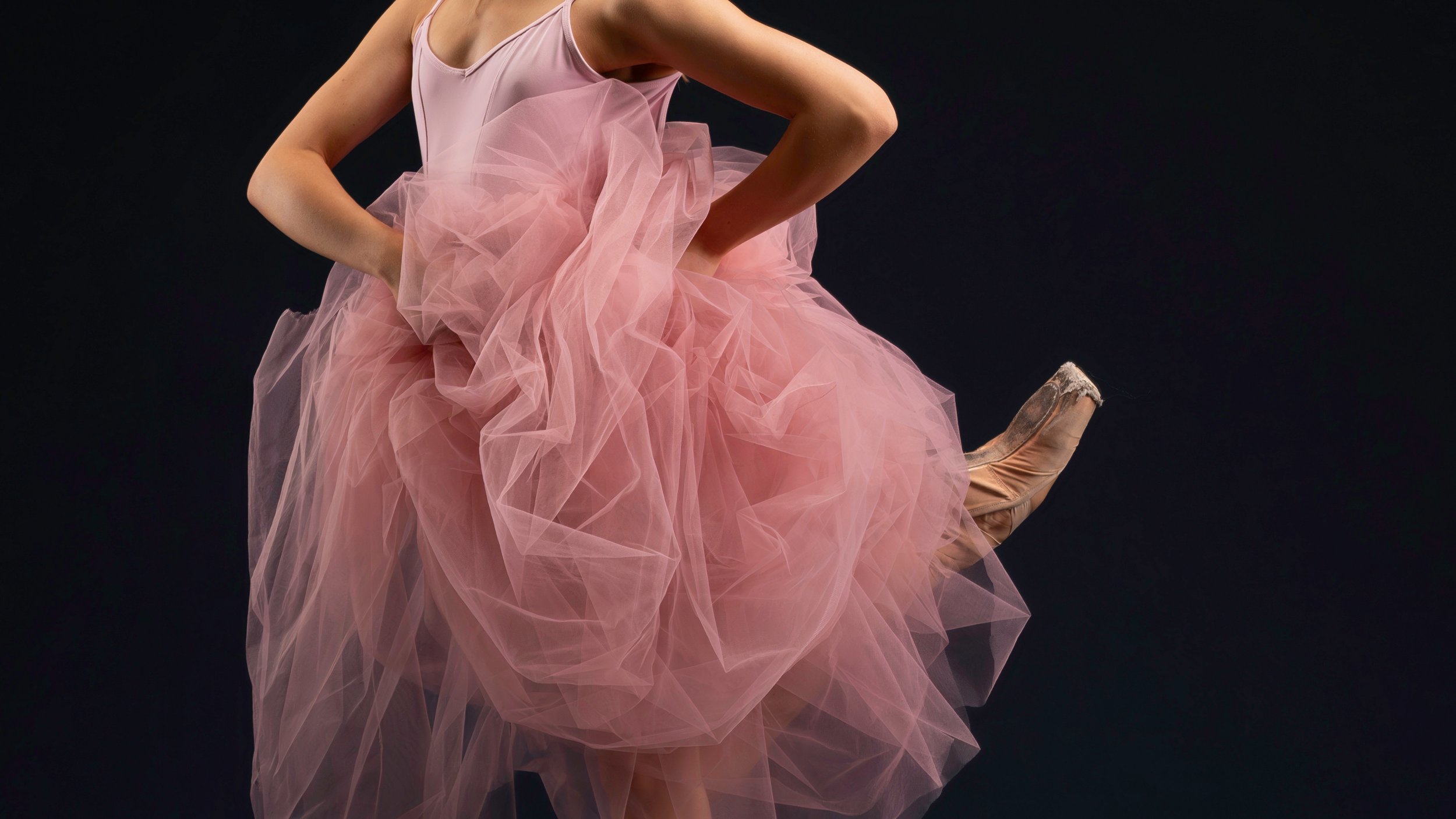 A ballet dancer wearing a pink tutu and pointe shoes, posed against a dark background.