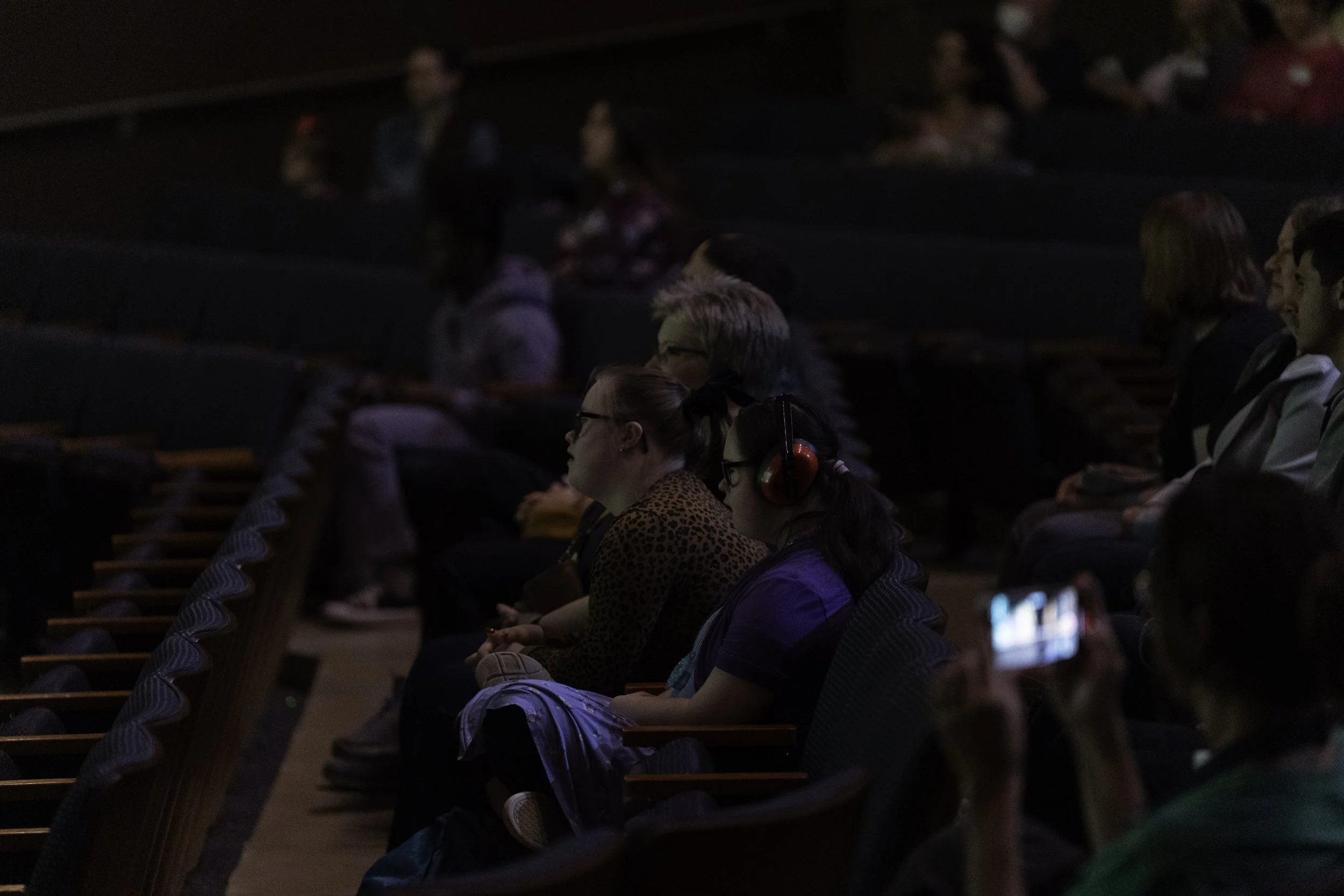 Audience members sitting in a darkened auditorium, some wearing headphones, watching a presentation or performance.
