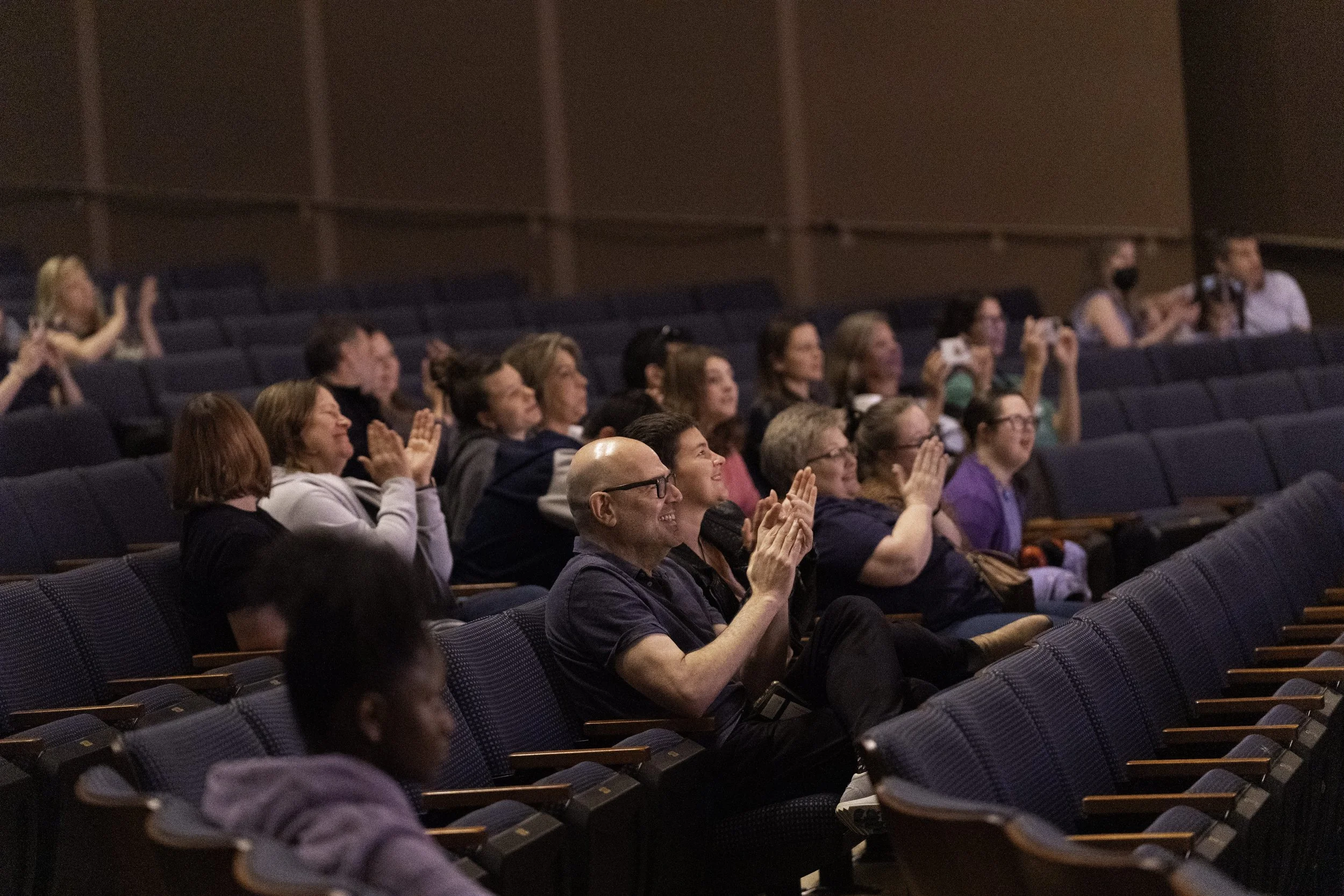 Audience members seated in a theater, clapping and smiling during a performance or presentation.