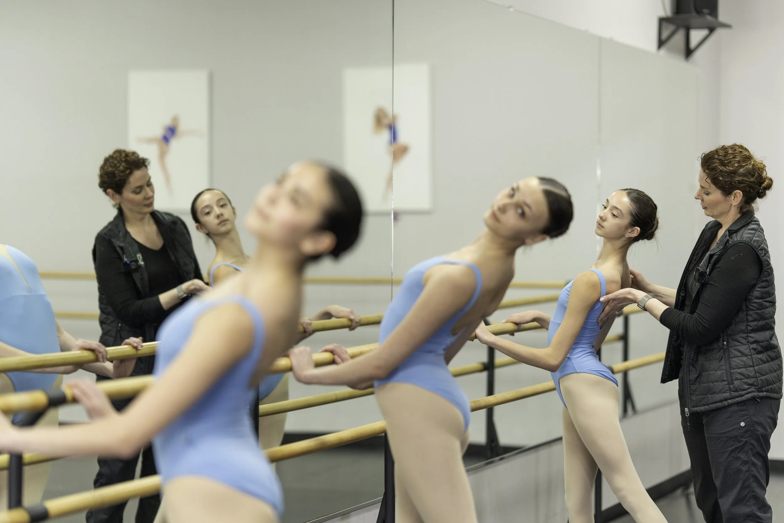 Ballet dancers in blue leotards practicing at the barre with an instructor adjusting a dancer's posture in a dance studio.