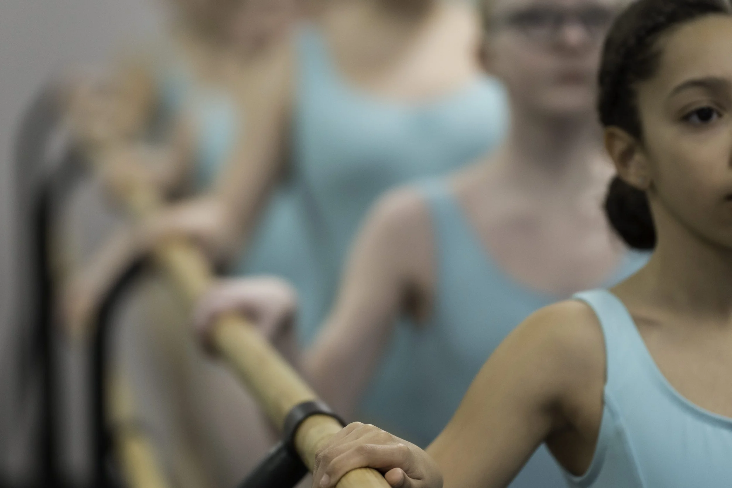 Three young girls in a ballet class wearing turquoise leotards, standing at the barre with one girl holding the barre with her hand.
