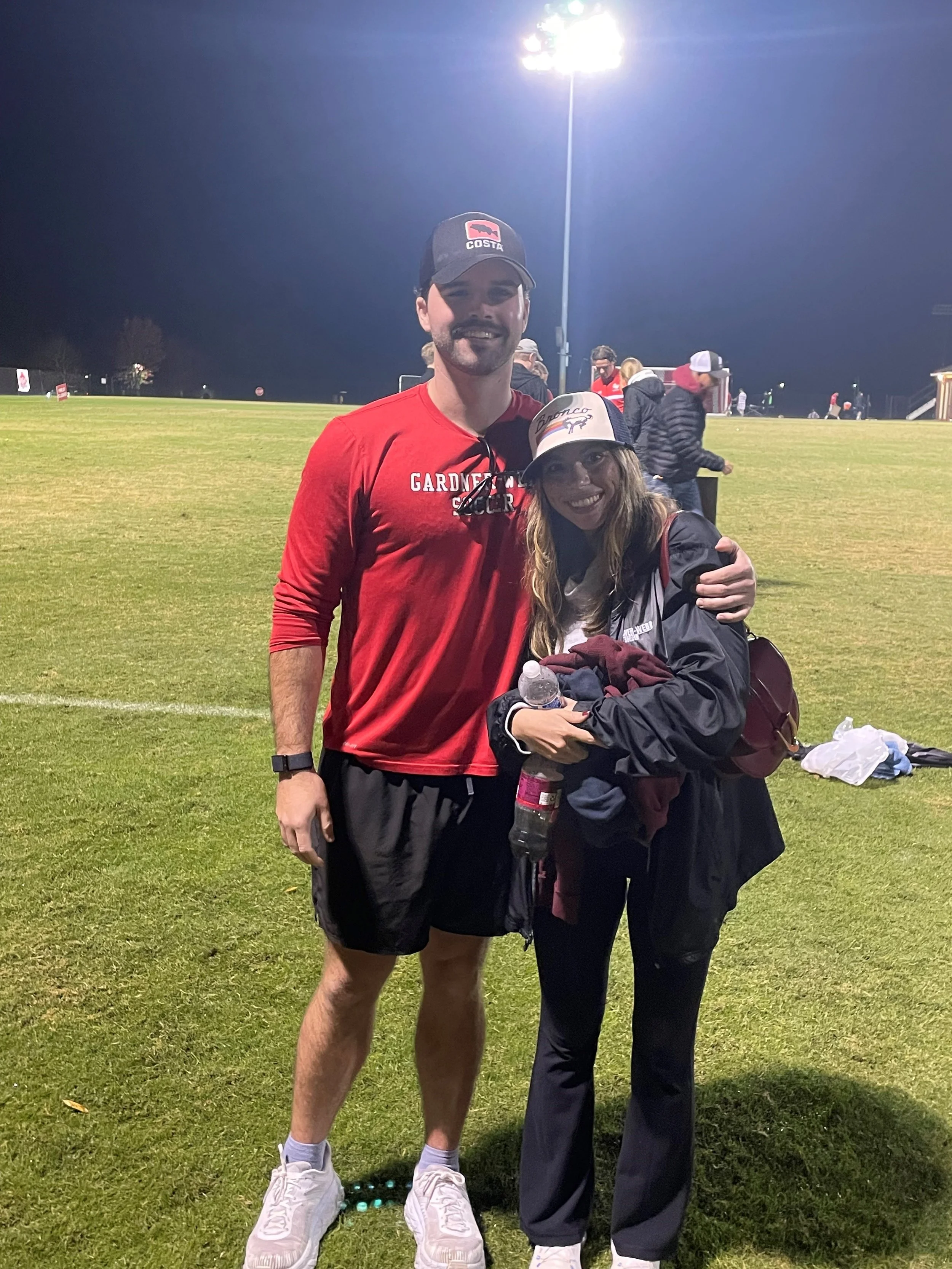 Two people standing on a sports field at night, one wearing a red GARDNER pitcher shirt and the other in a black jacket and baseball cap, smiling and posing for a photo.